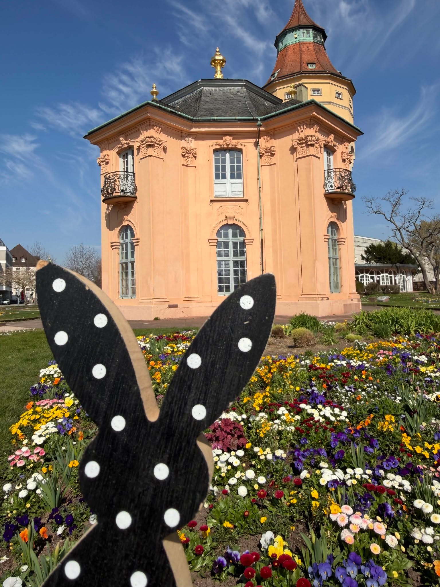 Dieser Alternativtext wurde KI-generiert: Ein Gebäude mit einem roten Turm steht inmitten eines farbenfrohen Blumengartens. Im Vordergrund befindet sich eine hölzerne Osterdekoration mit Polka-Dots.