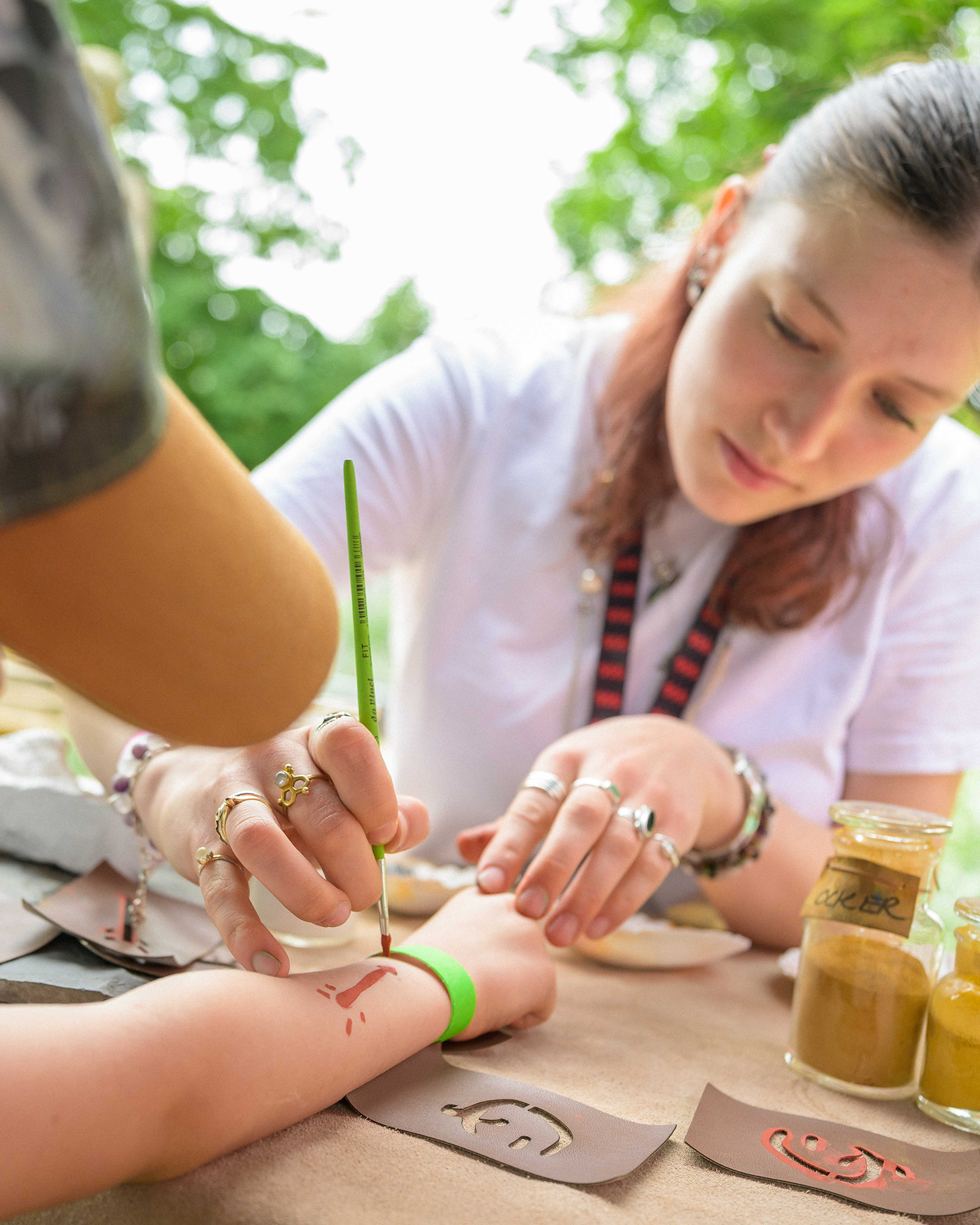 Eine Person malt mit einem grünen Pinsel ein rotes Muster auf den Unterarm einer anderen Person, die einen grünen Armband trägt. Auf dem Tisch liegen Schablonen und Gläser mit farbigen Pulvern, im Hintergrund sind grüne Bäume zu sehen. (KI-Alternativtext)
