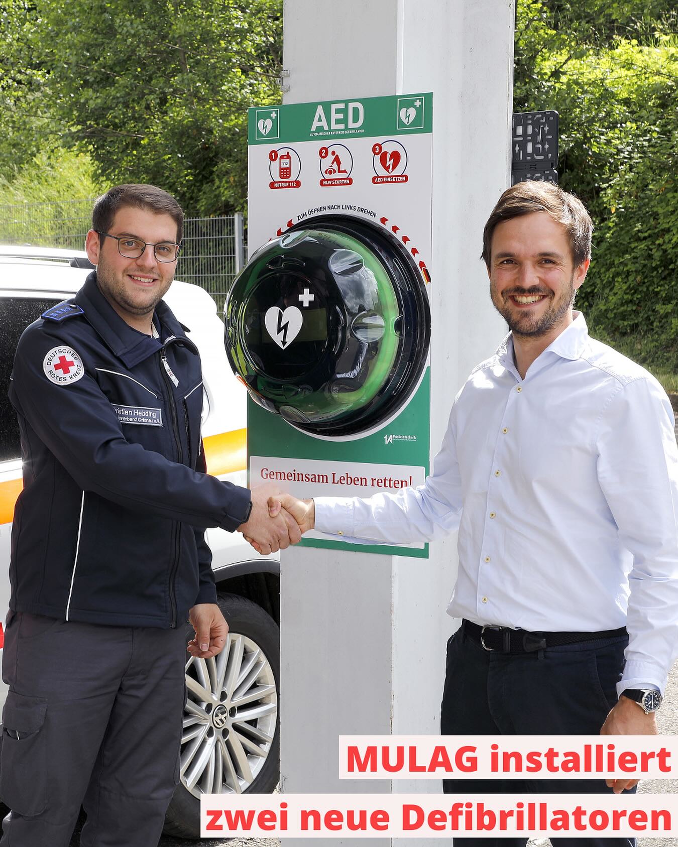 Zwei Personen geben sich vor einem Defibrillator die Hand. Im Hintergrund ist ein Fahrzeug zu sehen.