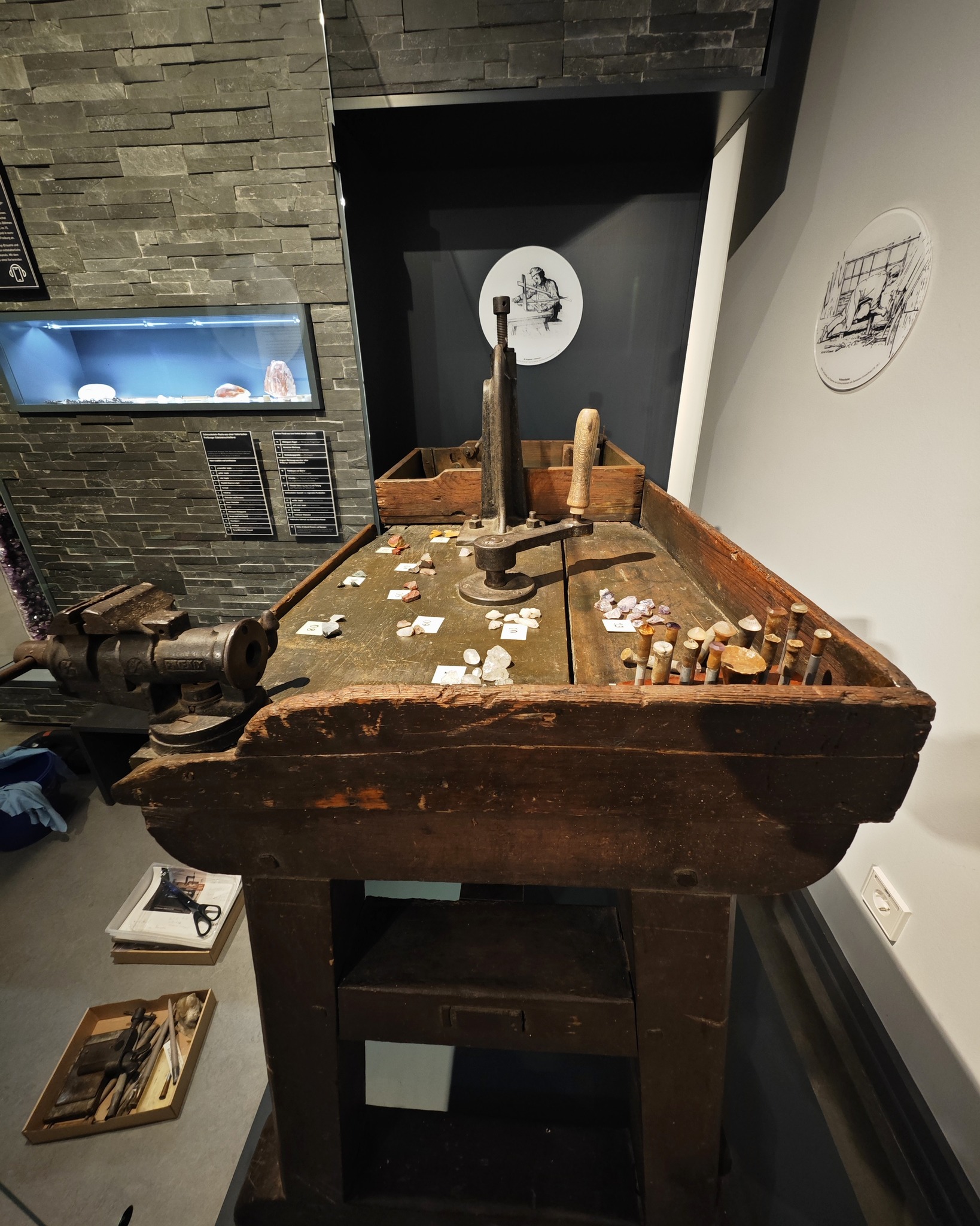 Ein alter, massiver Holzarbeitstisch mit einem Metallschraubstock, einer Presse und einer Reihe von Stichelwerkzeugen steht im Mittelpunkt einer Museumsausstellung. Auf der Tischplatte sind lose Steine und Kristalle mit kleinen Beschriftungskarten verteilt. Im Hintergrund sind eine beleuchtete Vitrine mit Mineralien, Informationsplakate und runde Illustrationen an der Wand zu sehen. (KI-Alternativtext)