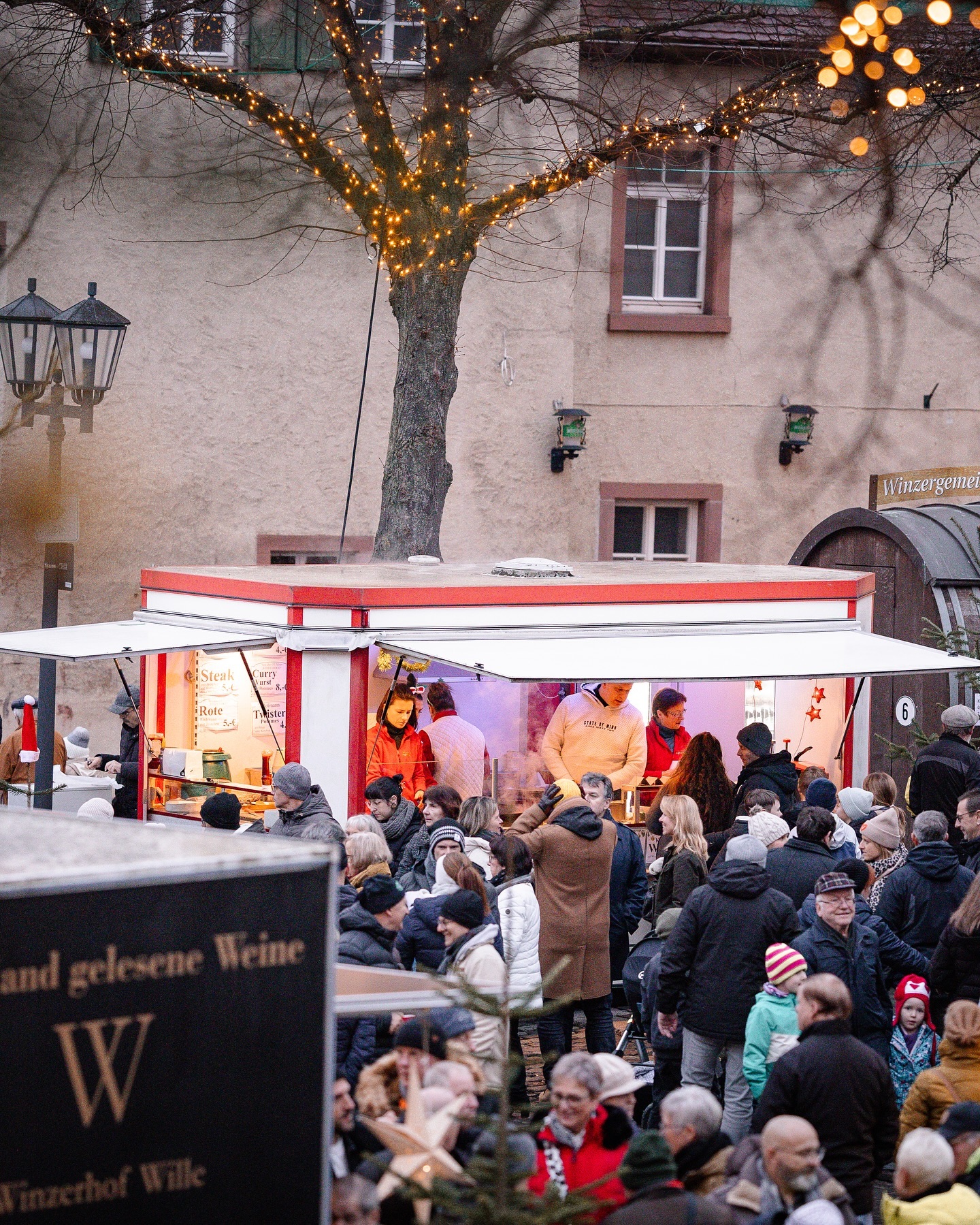 Ein Weihnachtsmarktstand mit vielen Menschen, die etwas kaufen. Ein Baum mit Lichtern steht über dem Stand.