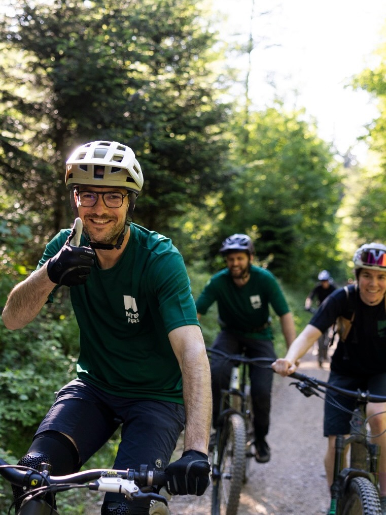Dieser Alternativtext wurde KI-generiert: Eine Gruppe von Menschen fährt auf einem Waldweg mit Mountainbikes. Die Person im Vordergrund trägt einen Helm und lächelt in die Kamera.