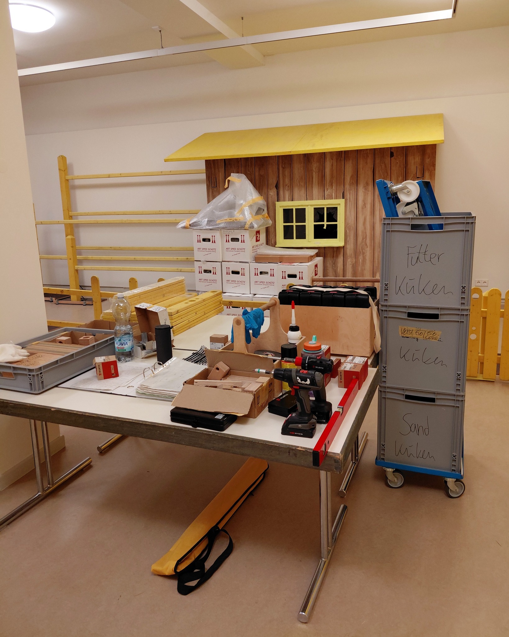 Dieser Alternativtext wurde KI-generiert: Ein Werkraum mit Werkzeugen und Baumaterialien. Auf einem Tisch befinden sich verschiedene Holzteile und Gegenstände, im Hintergrund ist eine halbfertige Holzkonstruktion zu sehen.