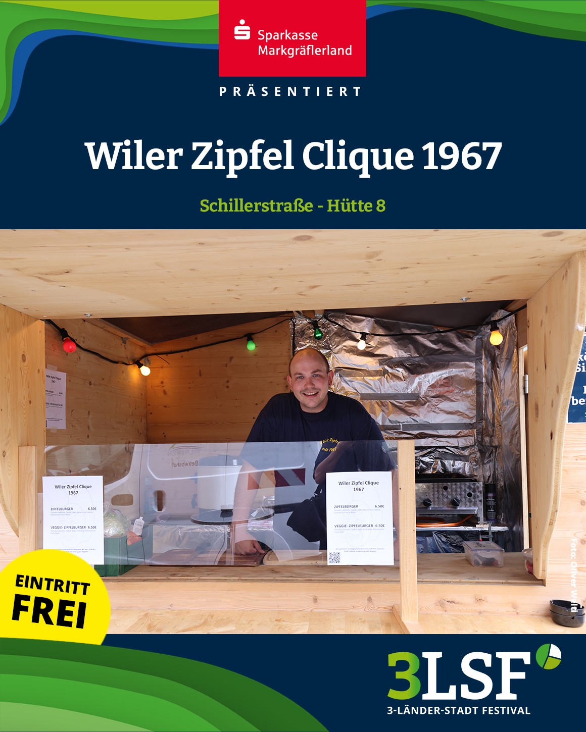 Dieser Alternativtext wurde KI-generiert: Ein Holzstand mit einer Person darin, die an einem Tisch sitzt. Im Vordergrund befindet sich ein gelber Werbeaufkleber mit der Aufschrift "EINTRITT FREI".