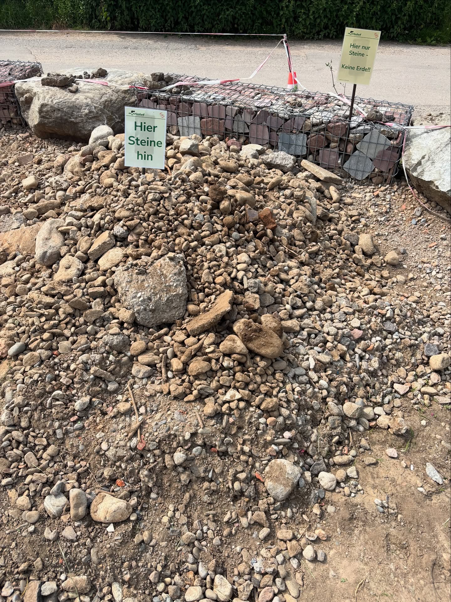 Ein Haufen Steine unterschiedlicher Größen liegt im Vordergrund. Im Hintergrund sind zwei Schilder und ein Drahtgitter zu sehen.
