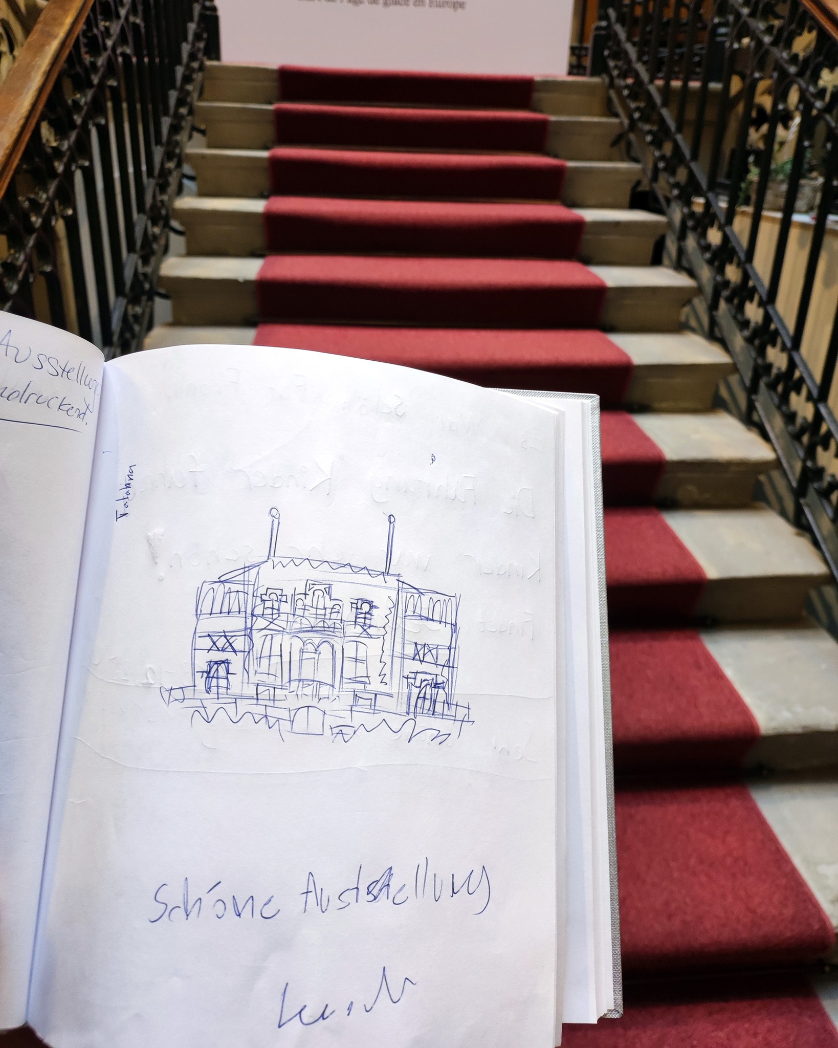 Dieser Alternativtext wurde KI-generiert: Ein offenes Buch mit einer Skizze eines Gebäudes liegt auf einer roten Treppe. Im Hintergrund ist ein helles Interieur mit einer Treppe zu sehen.