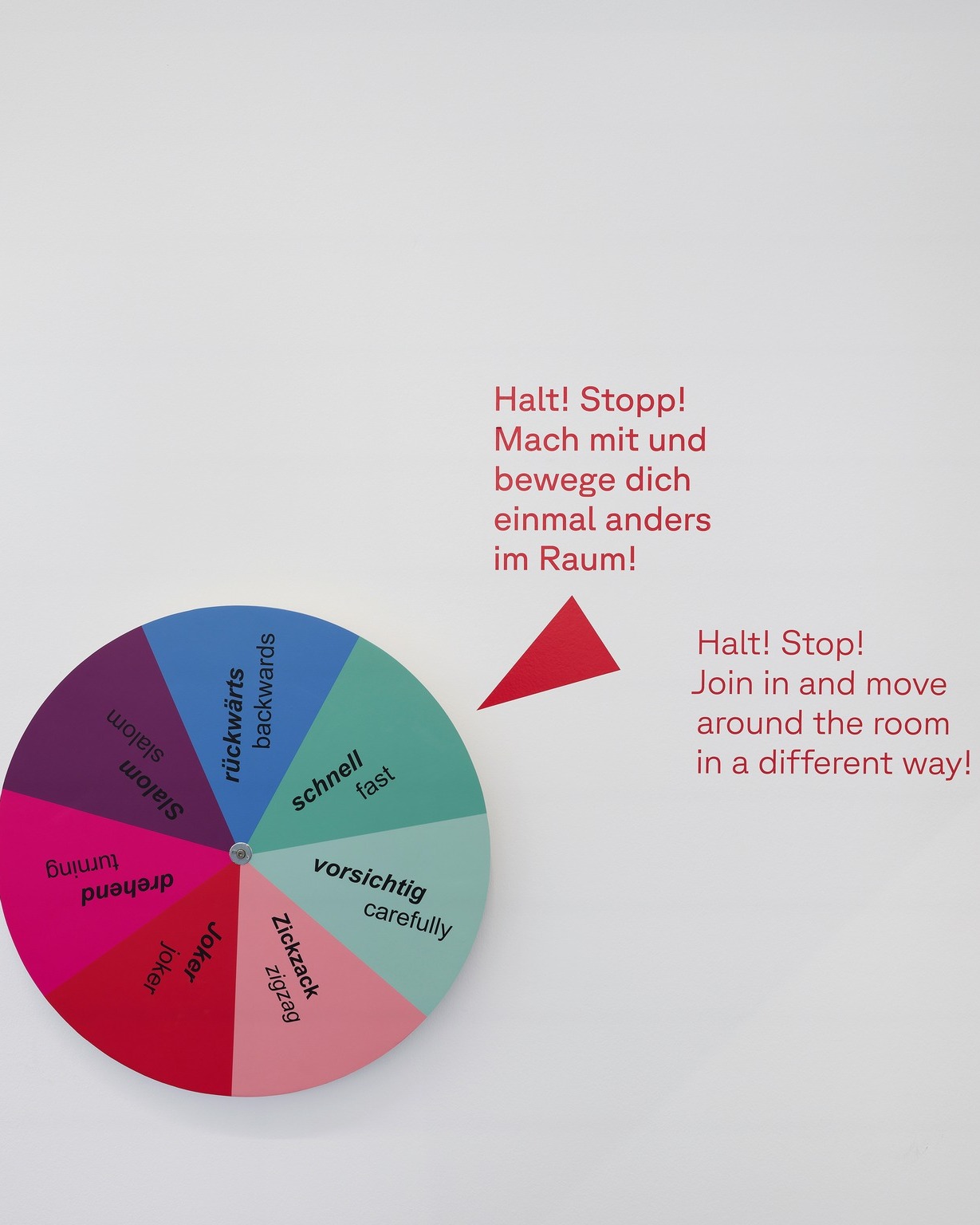 Ein farbiges Glücksrad mit sieben Segmenten trägt die Beschriftungen rückwärts, schnell, vorsichtig, Zickzack, Joker, drehend und Slalom, jeweils mit englischer Übersetzung. Rechts daneben steht in roter Schrift der Text „Halt! Stopp! Mach mit und bewege dich einmal anders im Raum!“ sowie die englische Version. Ein roter Pfeil zeigt auf das Rad. (KI-Alternativtext)