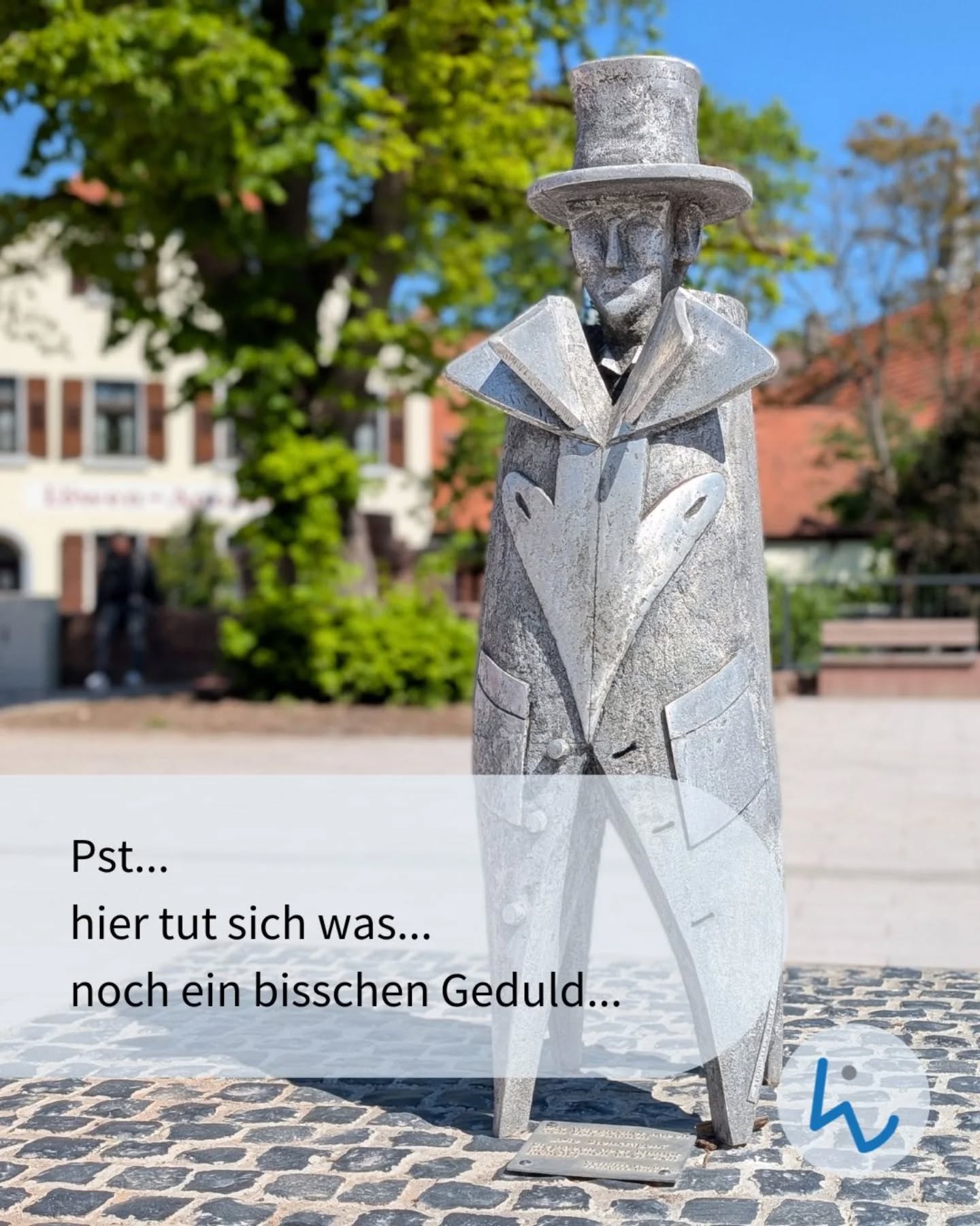 Dieser Alternativtext wurde KI-generiert: Eine graue Skulptur in formeller Kleidung steht auf einem gepflasterten Platz vor einem Gebäude. Ein Text über dem Bild lautet: "Pst... hier tut sich was... noch ein bisschen Geduld..."