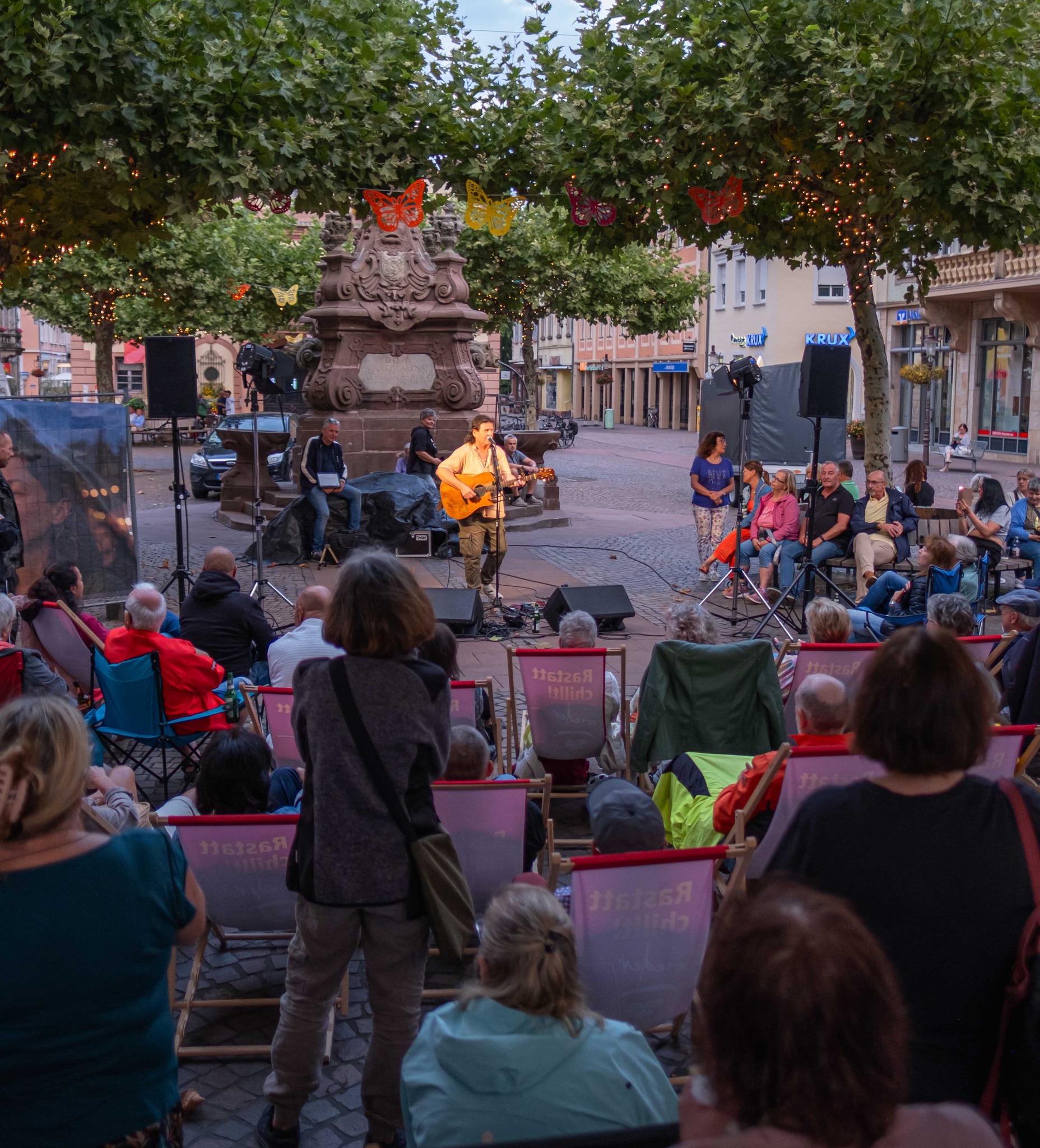 Eine Person spielt Gitarre und singt auf einer kleinen Bühne vor einem steinernen Denkmal. Das Publikum sitzt auf Klappstühlen mit der Aufschrift "Rastatt chillt!" und schaut zu. Über den Bäumen hängen Lichterketten und bunte Schmetterlingsdekorationen. (KI-Alternativtext)