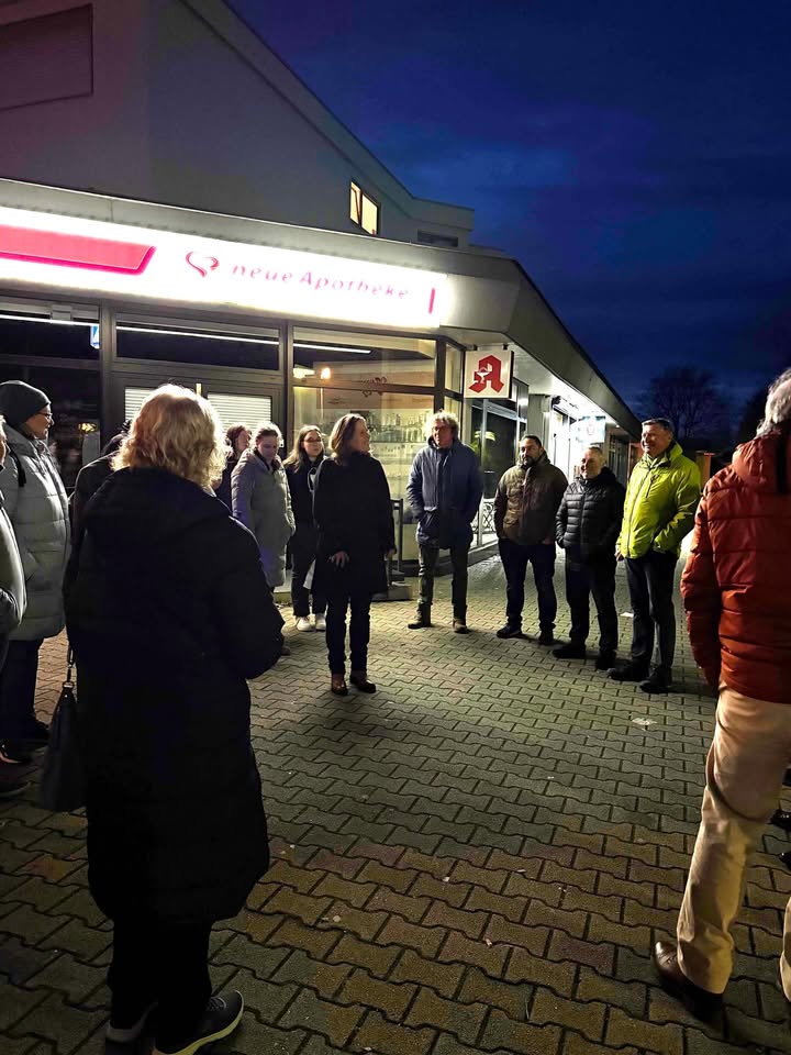 Dieser Alternativtext wurde KI-generiert: Eine Gruppe von Menschen steht vor einem Gebäude mit einer Apotheke. Es ist Nacht und die Szene ist teilweise im Schatten.