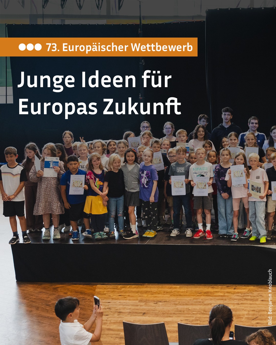 Gruppe von Kindern und Jugendlichen auf einer Bühne, die Zertifikate halten, vor einem Banner mit der Aufschrift „73. Europäischer Wettbewerb – Junge Ideen für Europas Zukunft“. Im Vordergrund fotografiert ein Kind die Gruppe. (KI-Alternativtext)