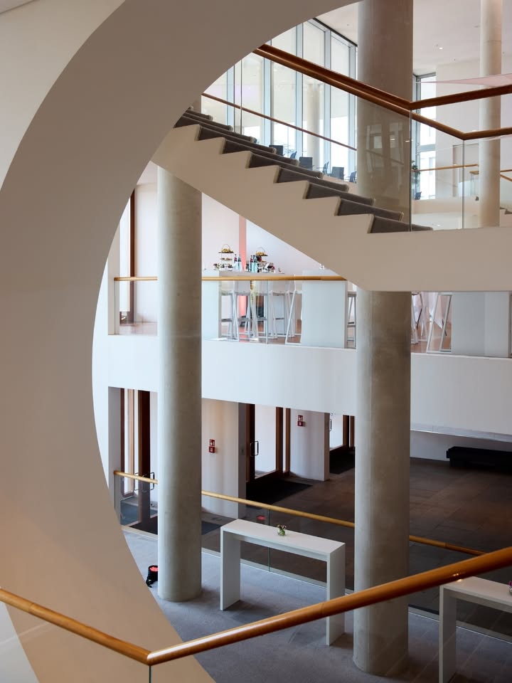 Durch eine große, kreisförmige Öffnung in einer weißen Wand blickt man in ein modernes, mehrstöckiges Gebäudeinnere. Massive Betonpfeiler stützen die Etagen, die durch Treppen mit grauen Stufen und Holzhandläufen sowie Glasgeländern verbunden sind. Im Vordergrund steht ein weißer Tisch, während auf der mittleren Ebene ein weiterer Tisch mit Gegenständen zu sehen ist. (KI-Alternativtext)