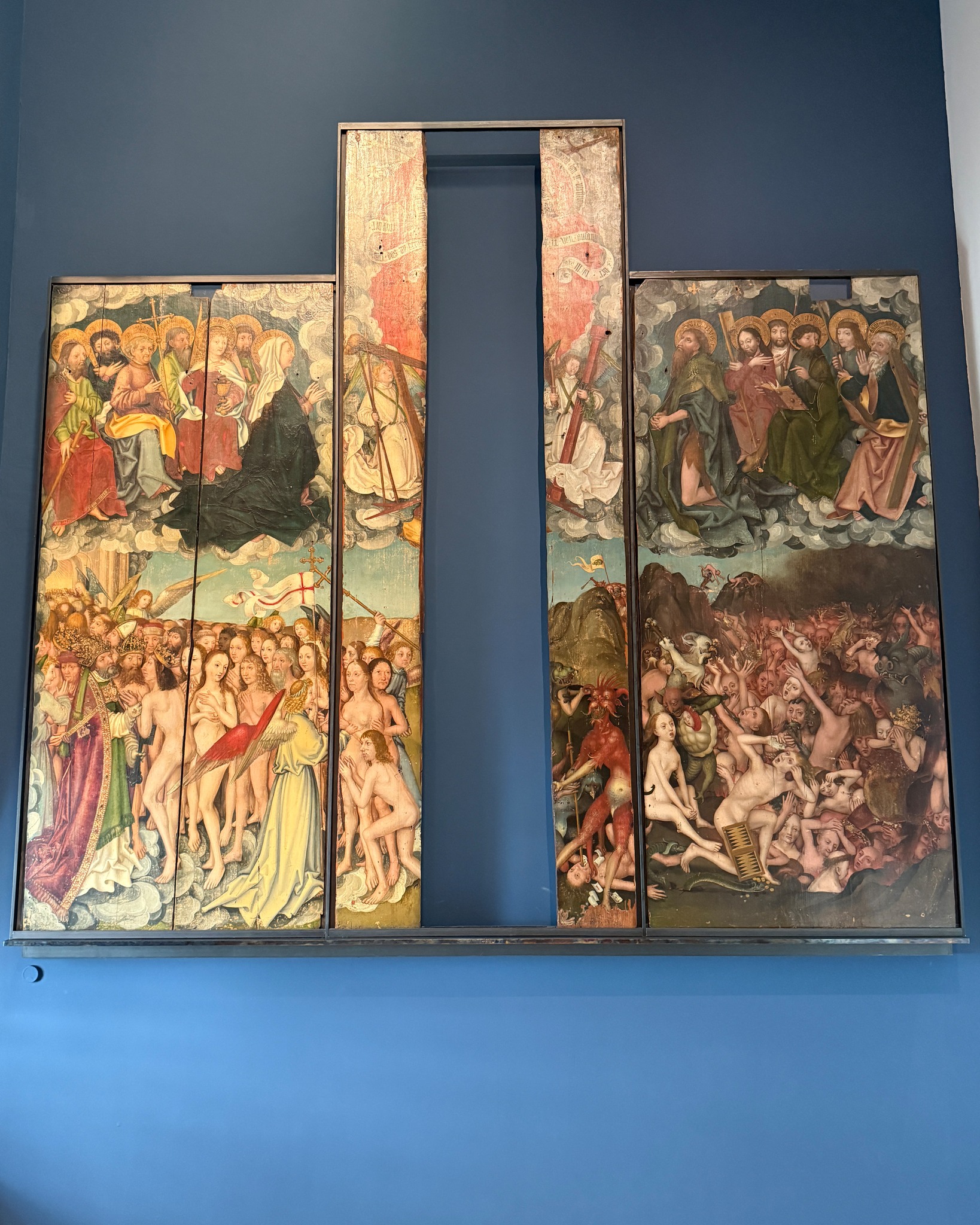 Ein großes, mehrteiliges Gemälde an einer blauen Wand, das das Jüngste Gericht darstellt. Auf der linken Seite sind selige Seelen und Heilige in den Wolken zu sehen, während die rechte Seite eine Hölle mit Dämonen und leidenden Figuren zeigt. In der Mitte trennen zwei schmale, vertikale Tafeln die Szenen voneinander. (KI-Alternativtext)