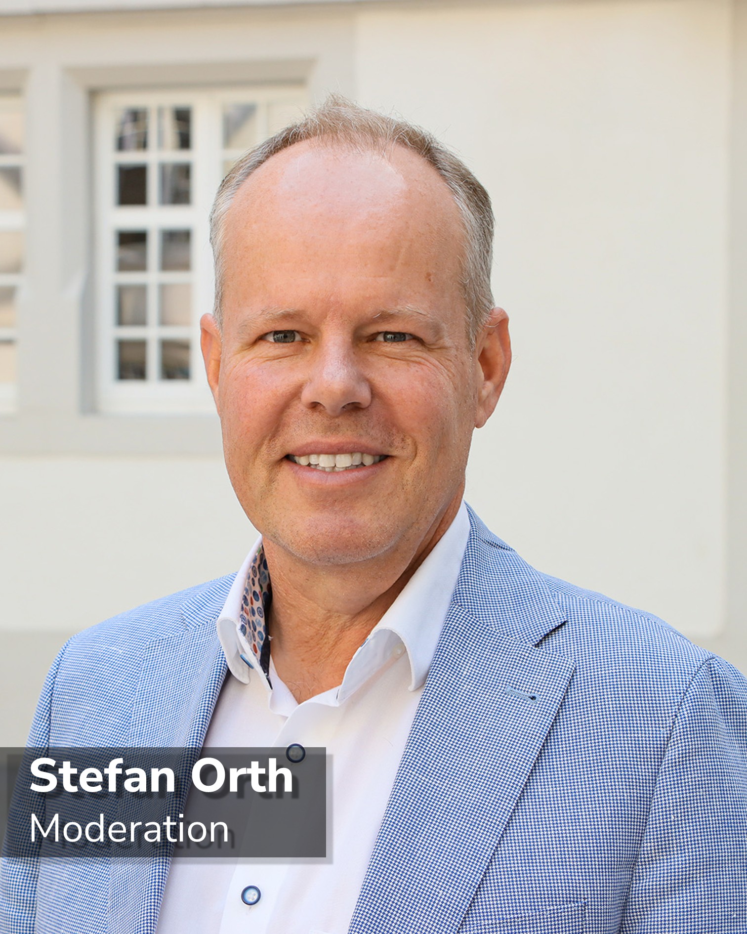 Dieser Alternativtext wurde KI-generiert: Ein Porträt einer Person, die lächelt und einen Blazer trägt. Im Hintergrund ist ein Gebäude mit Fenstern zu sehen.