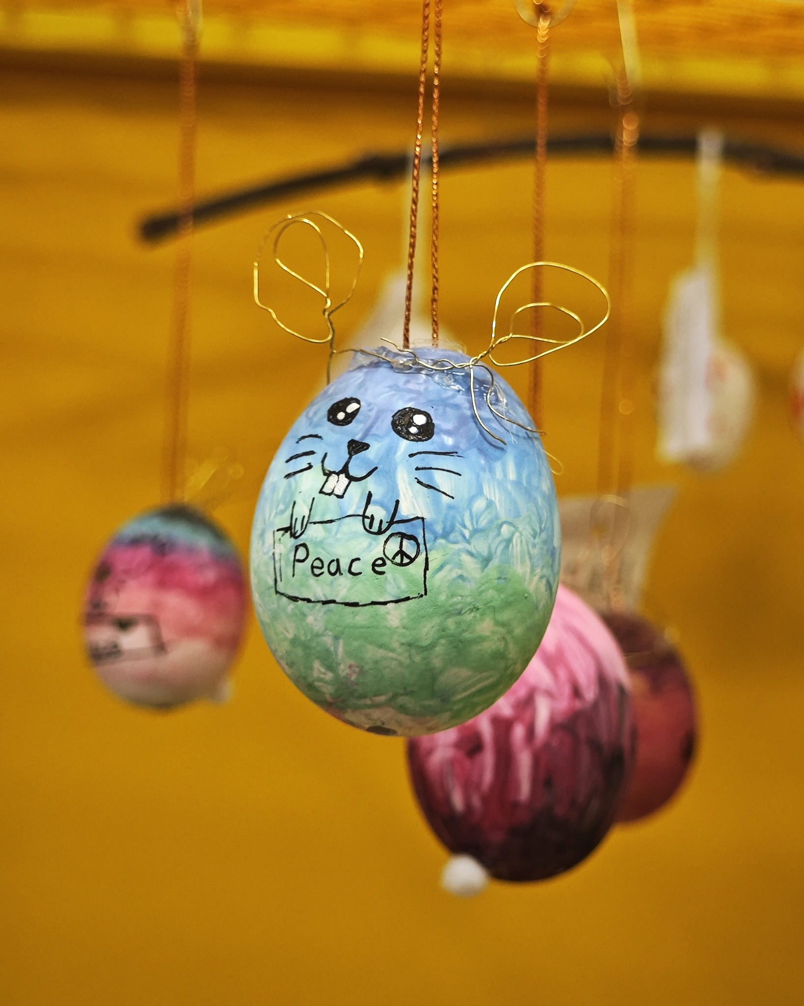 Dieser Alternativtext wurde KI-generiert: Mehrere bemalte Eier hängen an Schnüren vor einem gelben Hintergrund. Ein Ei ist blau-grün und mit einem Gesicht und einem Friedenszeichen versehen.