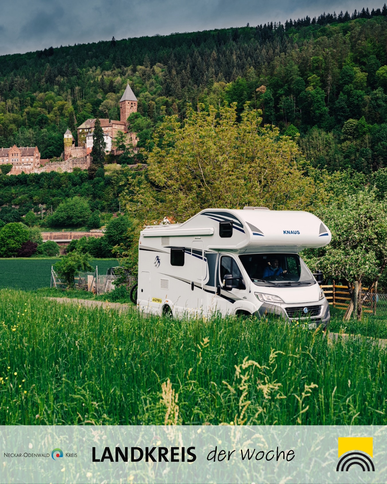 Dieser Alternativtext wurde KI-generiert: Ein Wohnmobil steht auf einer Wiese vor einer hügeligen Landschaft mit Bäumen und einem Gebäude auf einem Felsen. Das Bild ist farbig und zeigt eine ruhige Szene.