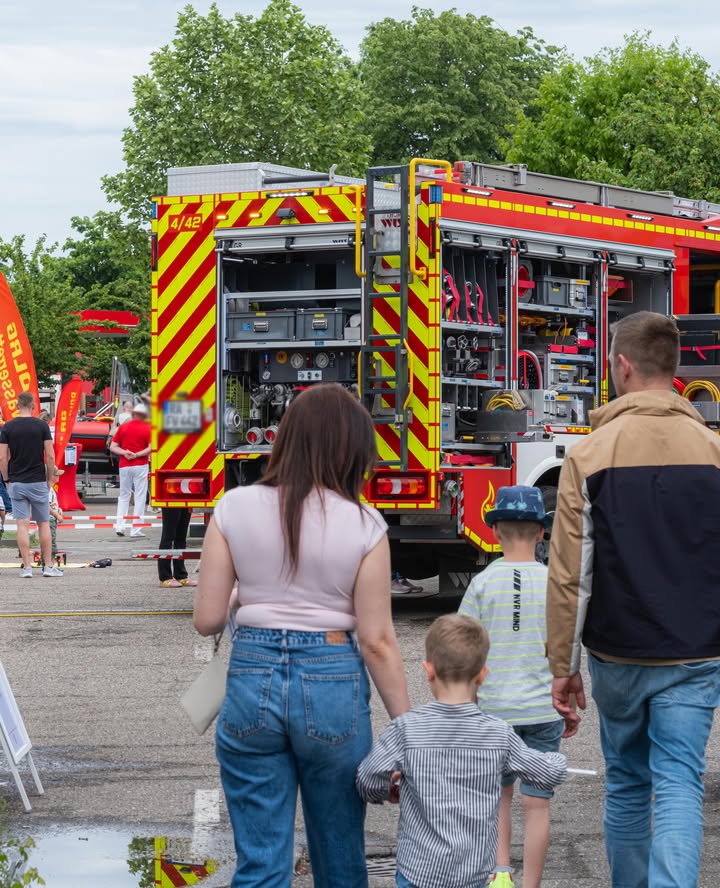 Dieser Alternativtext wurde KI-generiert: Ein rotes Feuerwehrauto steht im Vordergrund, betrachtet von mehreren Personen, die von hinten zu sehen sind. Die Szene spielt sich im Freien auf einer nassen Straße ab.