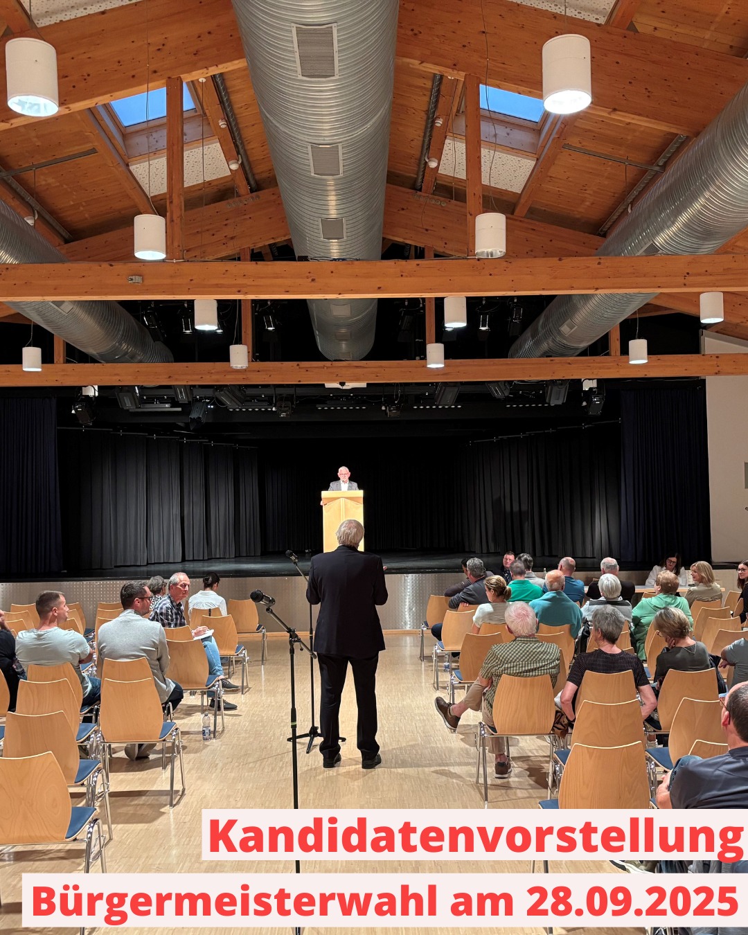 Dieser Alternativtext wurde KI-generiert: Ein großer Raum mit einer Bühne und Zuschauersitzen. Menschen sitzen und stehen, während eine Person auf der Bühne spricht, mit einem Banner im Vordergrund.