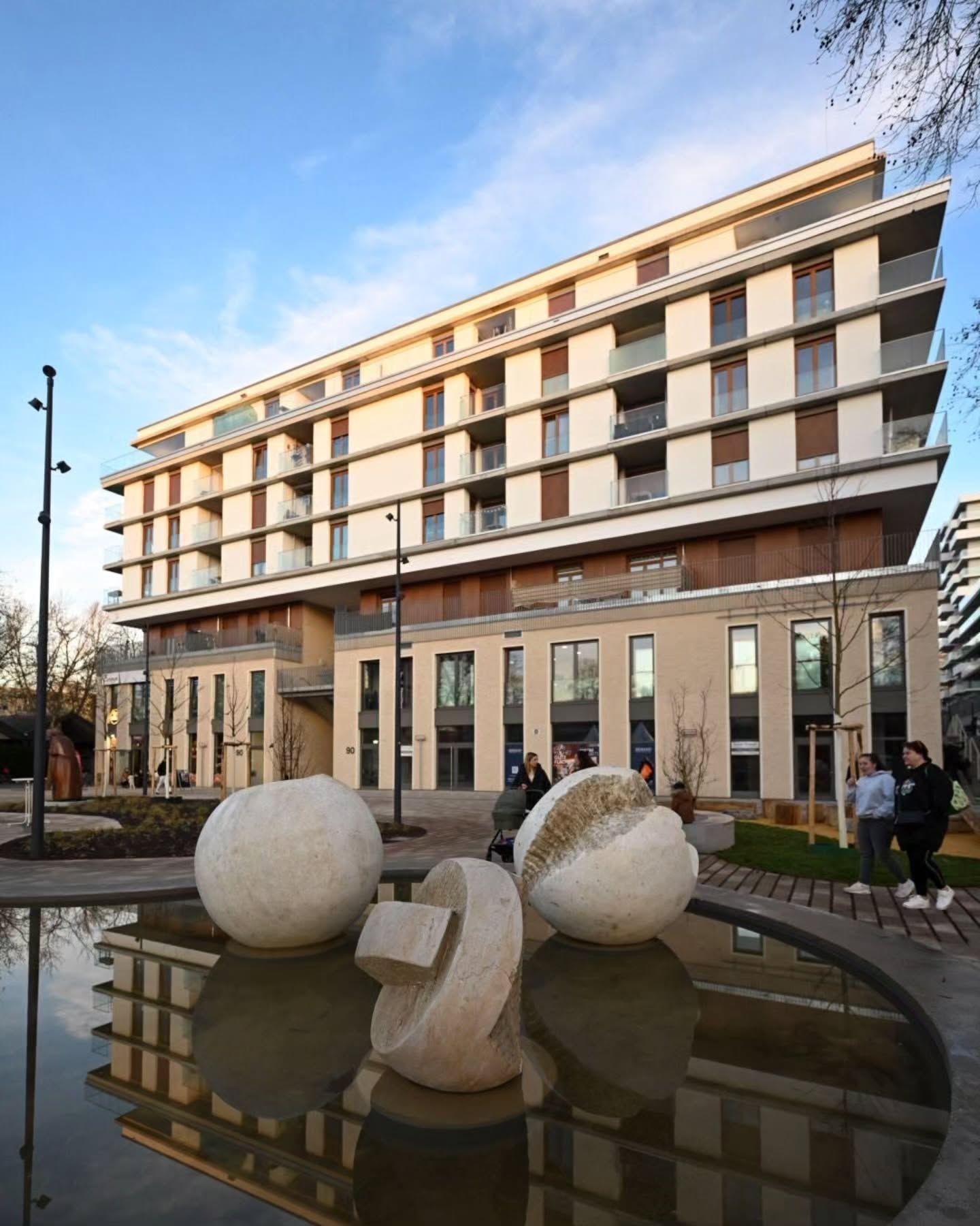 Dieser Alternativtext wurde KI-generiert: Ein modernes Gebäude mit mehreren Stockwerken steht im Vordergrund. Im Vordergrund befindet sich ein kleiner Platz mit Skulpturen und einem Wasserbecken.