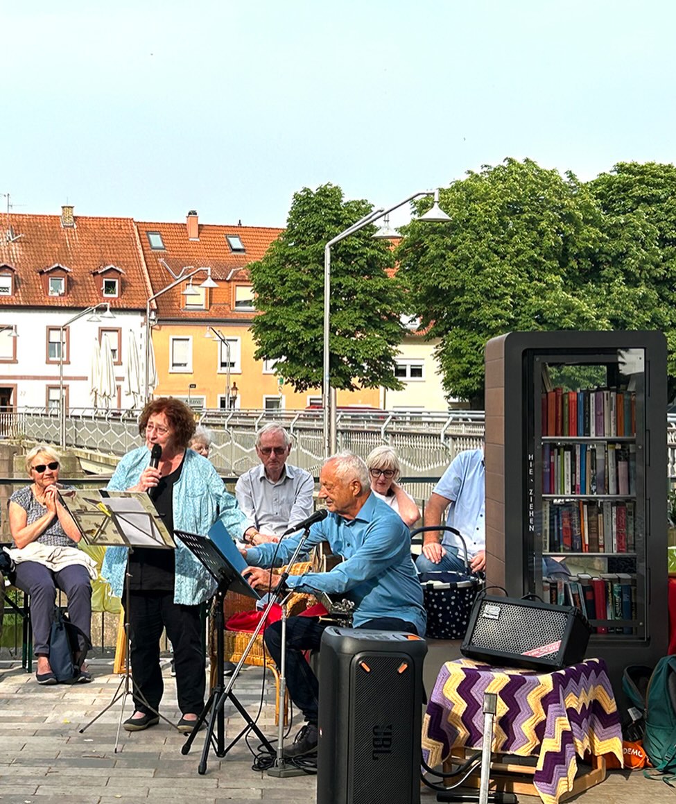 Eine Gruppe von Menschen musiziert im Freien, wobei eine Person singt und eine andere Gitarre spielt. Rechts steht ein schwarzes Bücherregal mit der Aufschrift 'HIER ZIEHEN' neben einem Tisch mit einer bunten Decke. Im Hintergrund sind Wohnhäuser und Bäume zu sehen. (KI-Alternativtext)