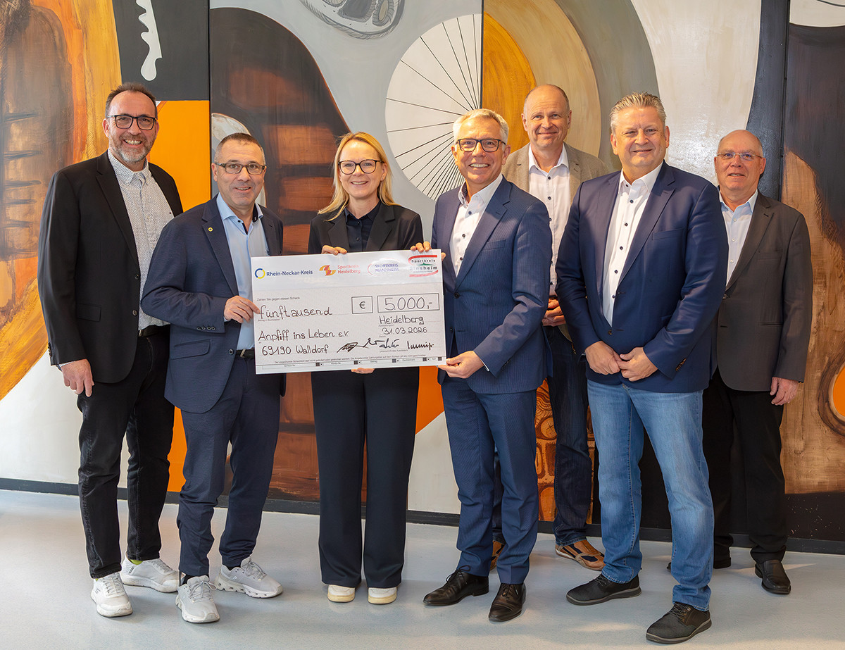 Dieser Alternativtext wurde KI-generiert: Eine Gruppe von Menschen in formeller Kleidung steht lächelnd vor einem Banner. Sie halten einen übergroßen Scheck in ihren Händen.