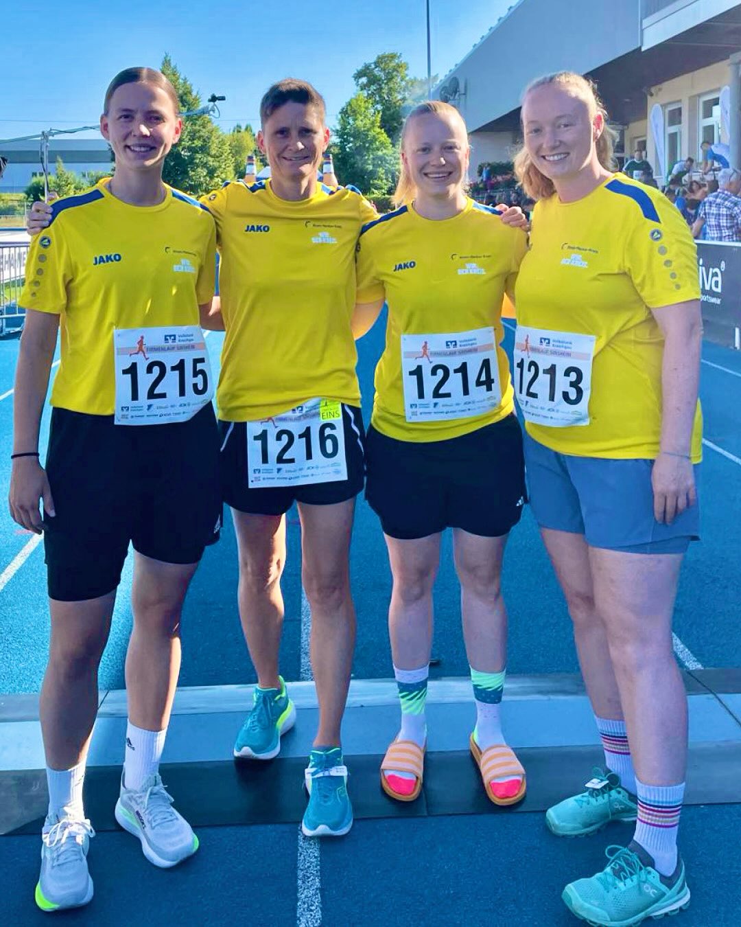 Vier Personen stehen mit Armen um die Schultern auf einer blauen Laufbahn und lächeln. Sie tragen gelbe Trikots mit dem Logo JAKO und Startnummern 1215, 1216, 1214 sowie 1213. Im Hintergrund sind Bäume und ein Gebäude zu sehen. (KI-Alternativtext)