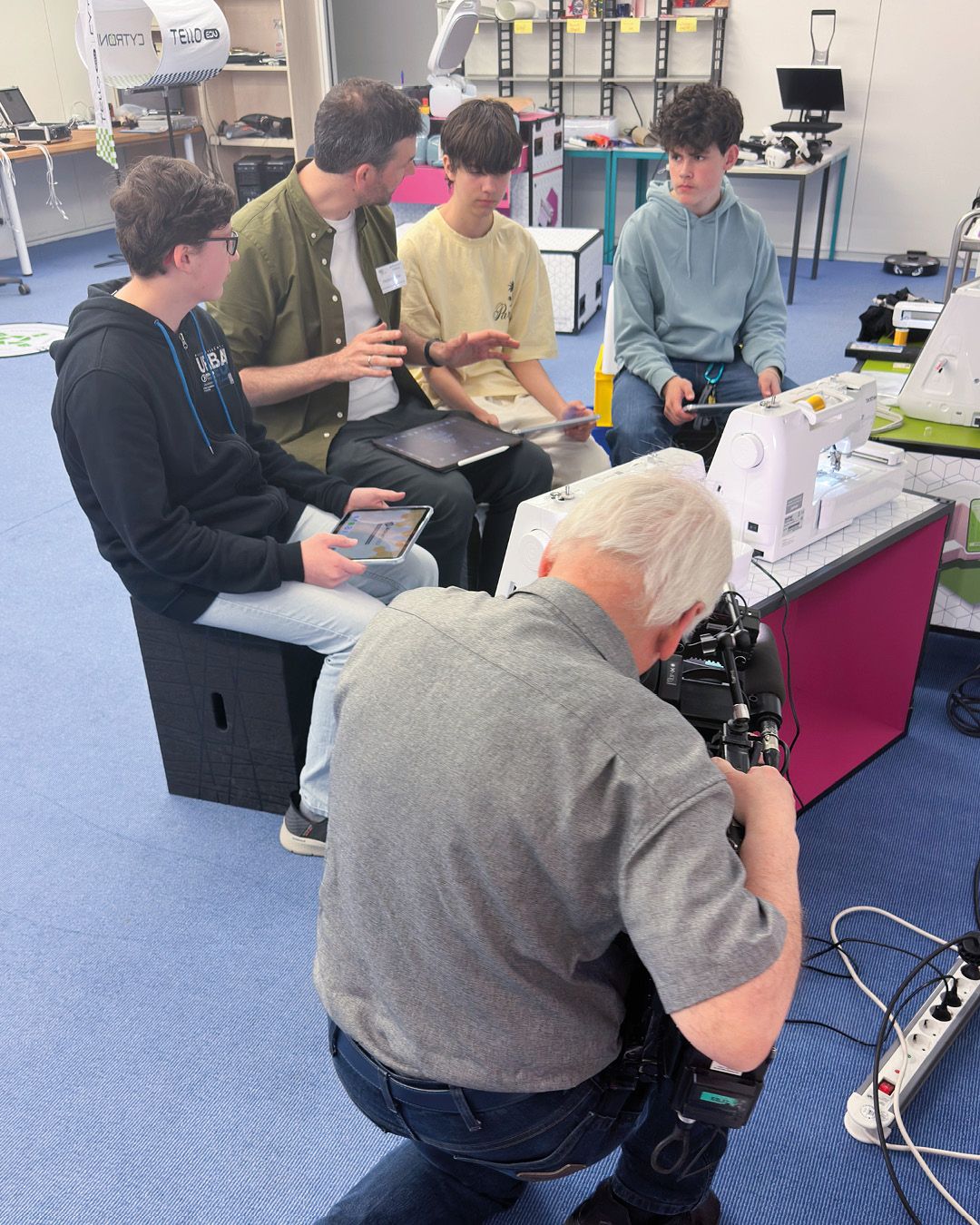 Dieser Alternativtext wurde KI-generiert: Eine Gruppe von Personen steht und sitzt in einem Raum mit technischer Ausrüstung. Eine Person filmt mit einer Kamera, während andere sich unterhalten und Geräte bedienen.