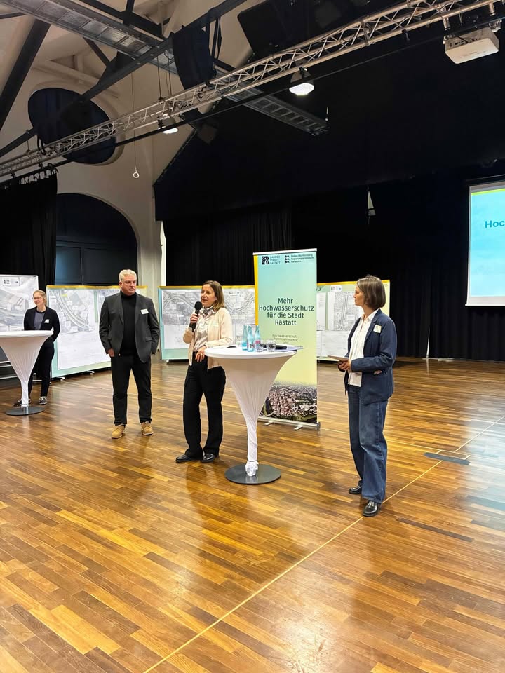 Dieser Alternativtext wurde KI-generiert: Eine Gruppe von Personen steht auf einer Bühne und vor einem Podest. Im Hintergrund ist ein Banner zu sehen und die Aufnahme ist farbig.