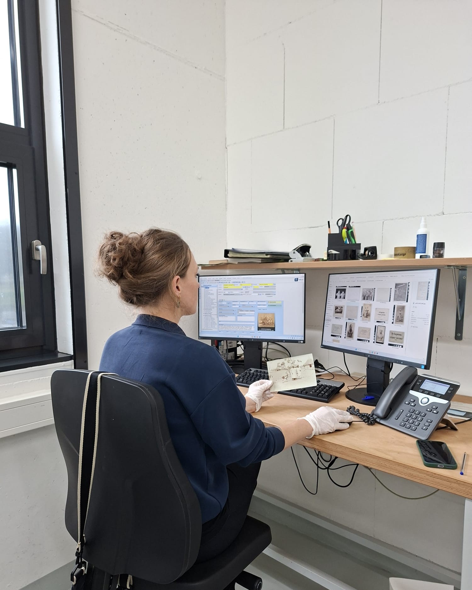 Dieser Alternativtext wurde KI-generiert: Eine Person sitzt an einem Schreibtisch vor zwei Computerbildschirmen und arbeitet. Auf dem Schreibtisch befinden sich verschiedene Gegenstände, darunter eine Telefonanlage und eine alte Fotografie.