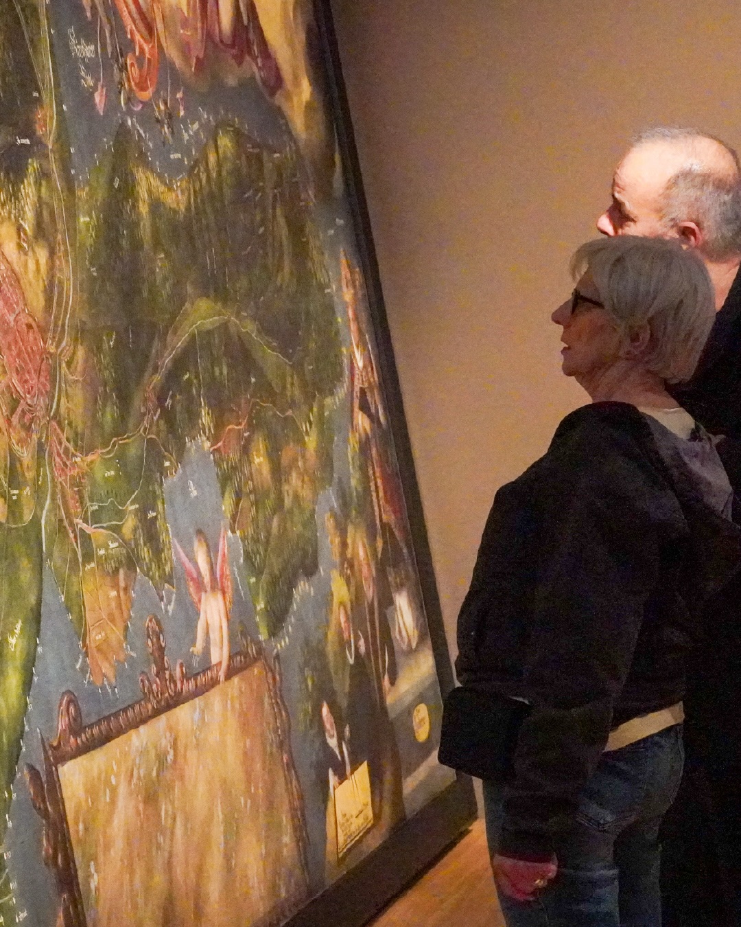 Dieser Alternativtext wurde KI-generiert: Zwei Personen betrachten ein großes, farbenfrohes Gemälde in einem Innenraum. Das Bild zeigt eine Landschaft mit abstrakten Elementen und menschlichen Figuren.