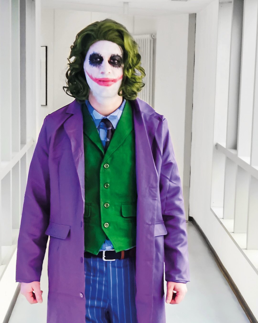 Dieser Alternativtext wurde KI-generiert: Eine Person trägt ein grünes Haarperücke und auffälliges Make-up. Sie ist mit einem lila Anzug und einer grünen Weste bekleidet und steht in einem hellen Flur.