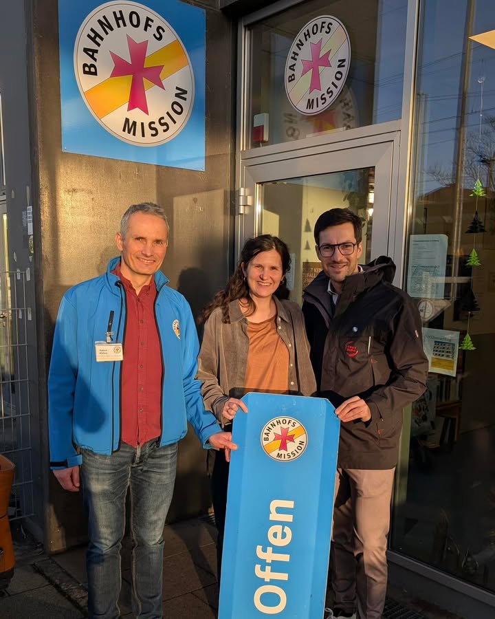 Dieser Alternativtext wurde KI-generiert: Drei Personen stehen vor einem Eingang mit Glasfront und halten ein Schild mit einem Logo. Das Bild ist farbig und zeigt eine Außenansicht mit metallischem Geländer.