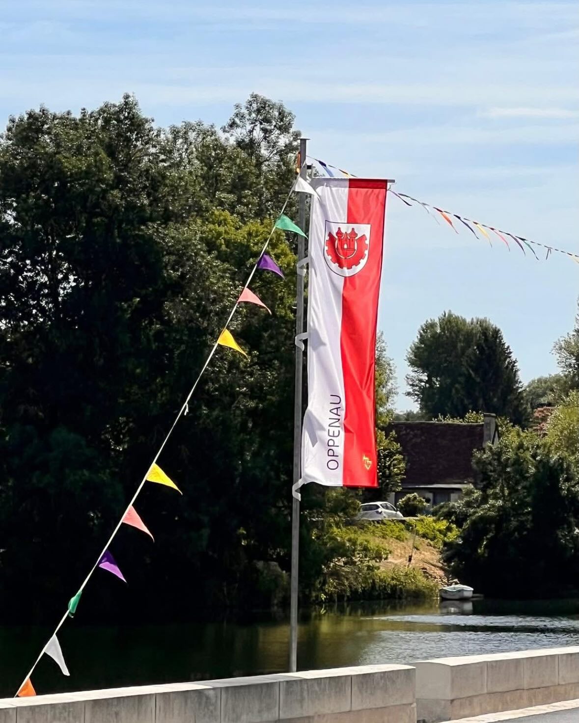 Eine rot-weiße Flagge hängt an einem Metallmast, daneben eine bunte Wimpelkette. Im Hintergrund sind ein Gewässer, diverse Bäume und ein Gebäude zu sehen.