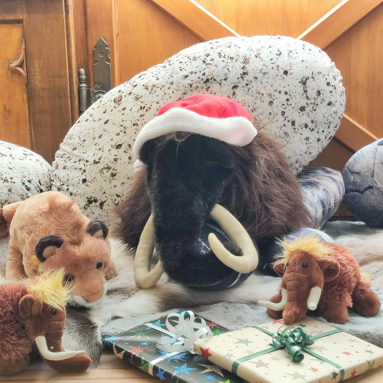 Dieser Alternativtext wurde KI-generiert: Mehrere Stofftiere, darunter ein wolliges Tier mit einem Weihnachtshut, liegen zusammen mit einem Geschenk auf einem Boden. Die Szene wirkt festlich und verspielt.