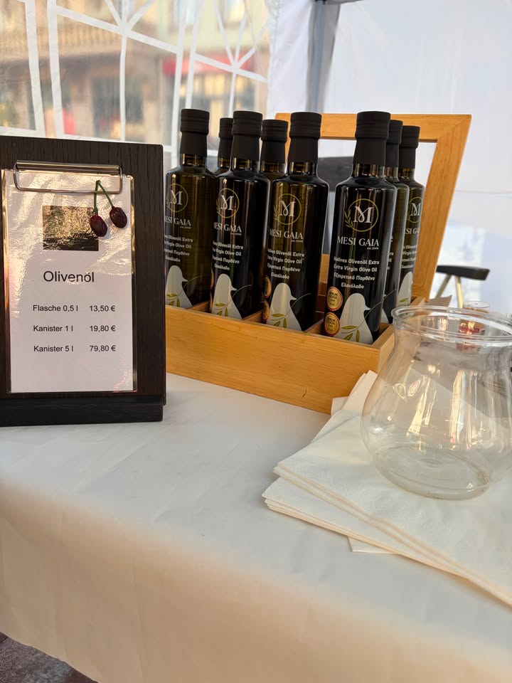 Ein Verkaufsstand mit Olivenöl. Links steht ein Schild mit der Aufschrift "Olivenöl" und den Preisen: Flasche 0,5 l für 13,50 €, Kanister 1 l für 19,80 € und Kanister 5 l für 79,80 €. Daneben stehen mehrere dunkle Flaschen der Marke Mesi Gaia in einem Holzkasten, rechts liegt ein Glasbehälter auf Servietten. (KI-Alternativtext)