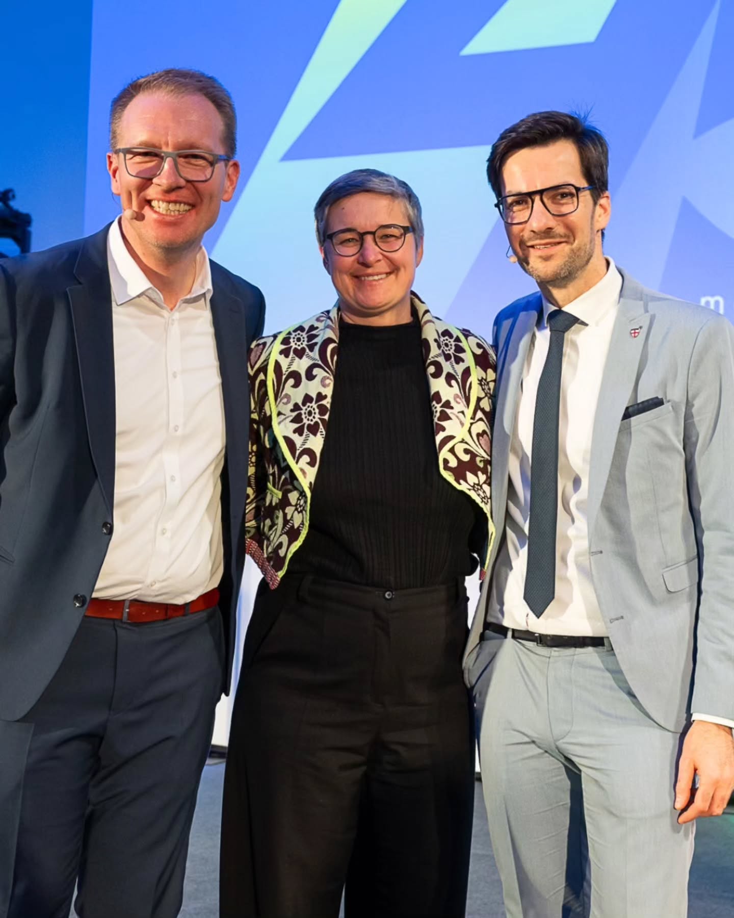 Dieser Alternativtext wurde KI-generiert: Drei Personen stehen lächelnd vor einem blauen Hintergrund. Alle tragen formelle Kleidung und Brillen.