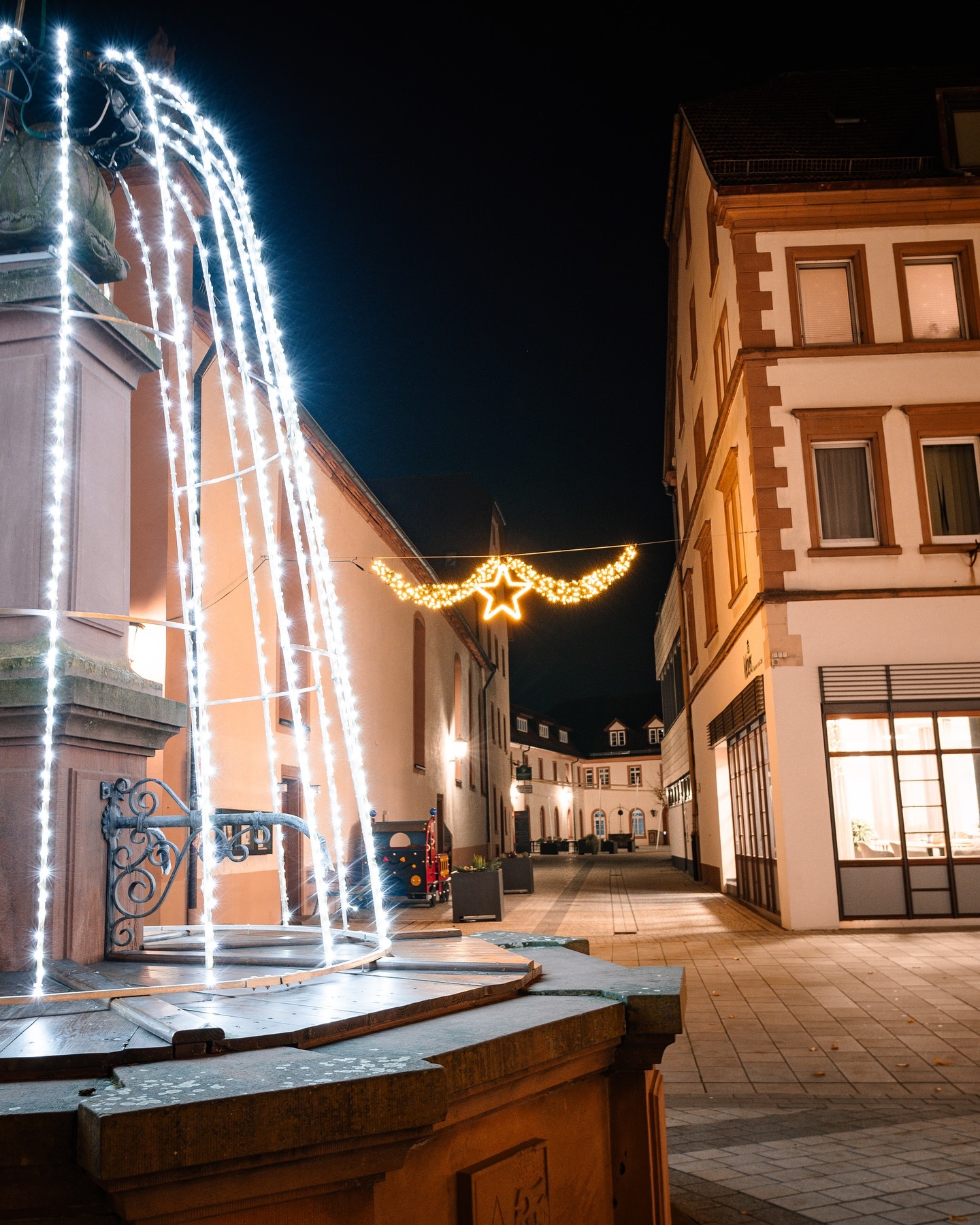 Ein Brunnen mit Lichterkette an einem Abend beleuchtet. Im Hintergrund sind Gebäude zu sehen, die ebenfalls weihnachtlich dekoriert sind.