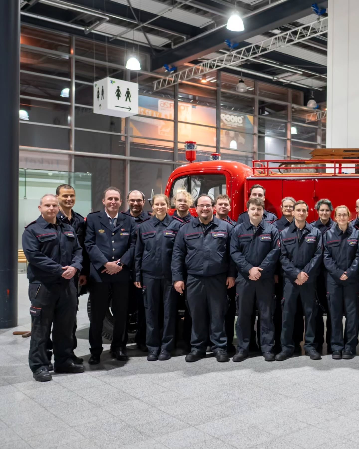 Dieser Alternativtext wurde KI-generiert: Eine Gruppe von Personen in dunkler Arbeitskleidung steht vor einem roten Feuerwehrauto in einer hellen Halle. Im Hintergrund sind weitere Personen und Schilder zu sehen.