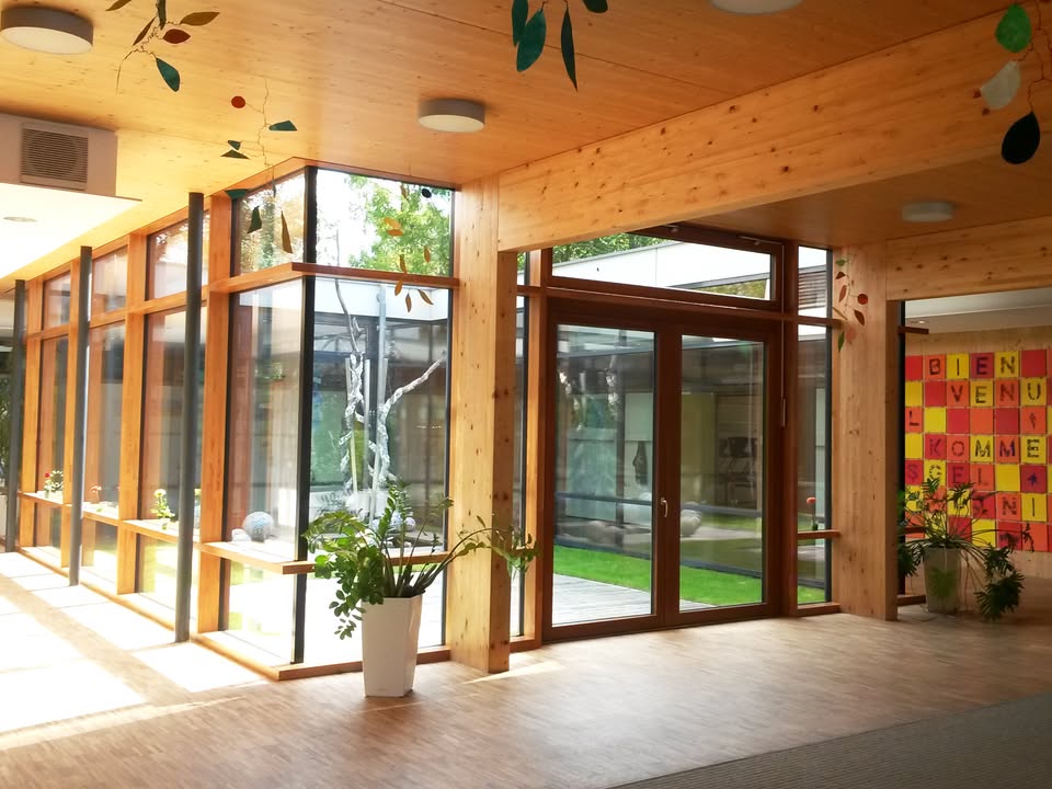 Dieser Alternativtext wurde KI-generiert: Ein heller Innenraum mit viel Holz und großen Fenstern, die einen Blick auf einen Garten ermöglichen. Eine Pflanze steht im Vordergrund.