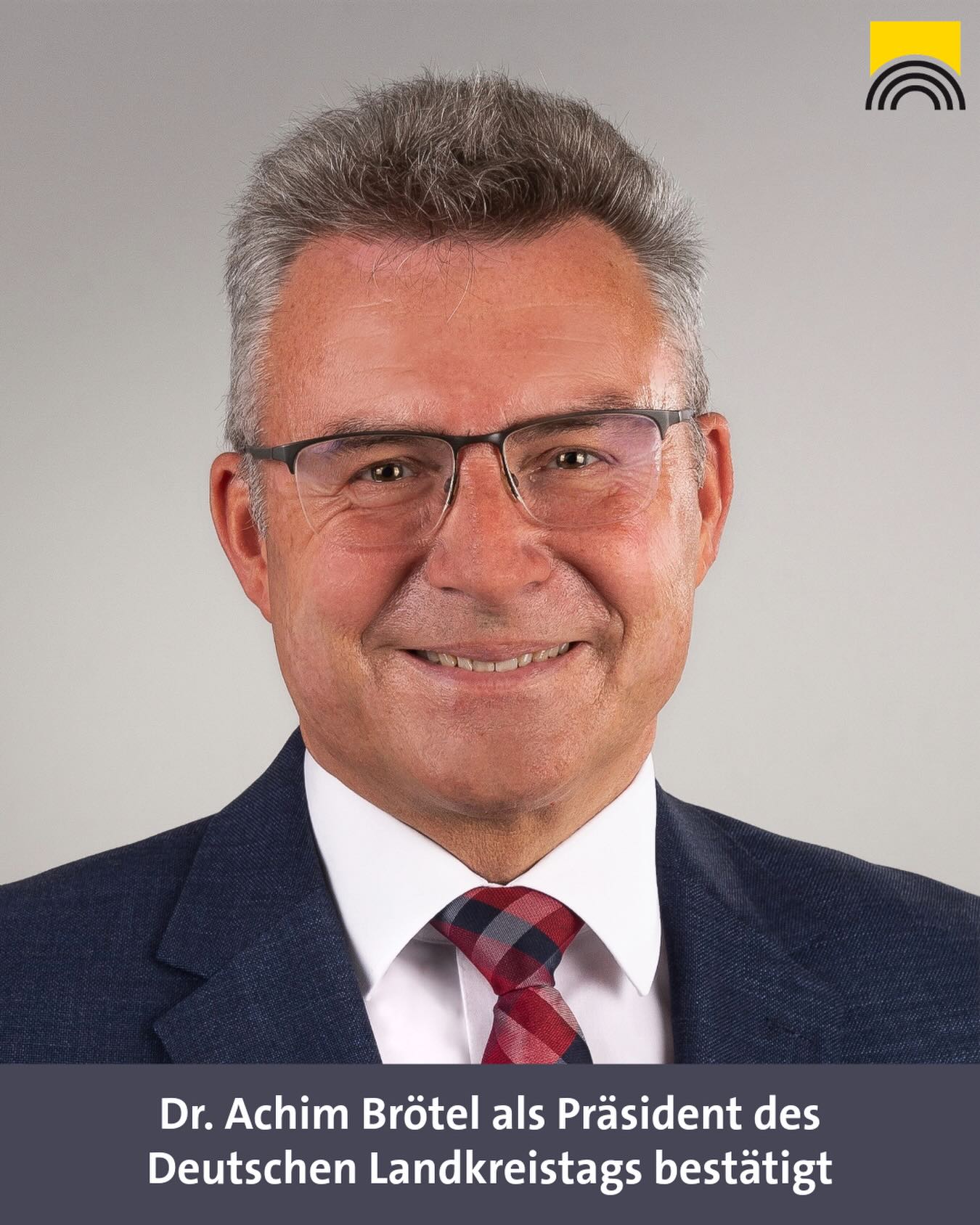 Menschen mit grauem Haar und Brille, in Anzug mit kariertem Krawatte, lächelt vor grauem Hintergrund. Unten steht: „Dr. Achim Brötel als Präsident des Deutschen Landkreistags bestätigt“. Oben rechts Logo mit gelbem Quadrat und schwarzen Bögen. (KI-Alternativtext)