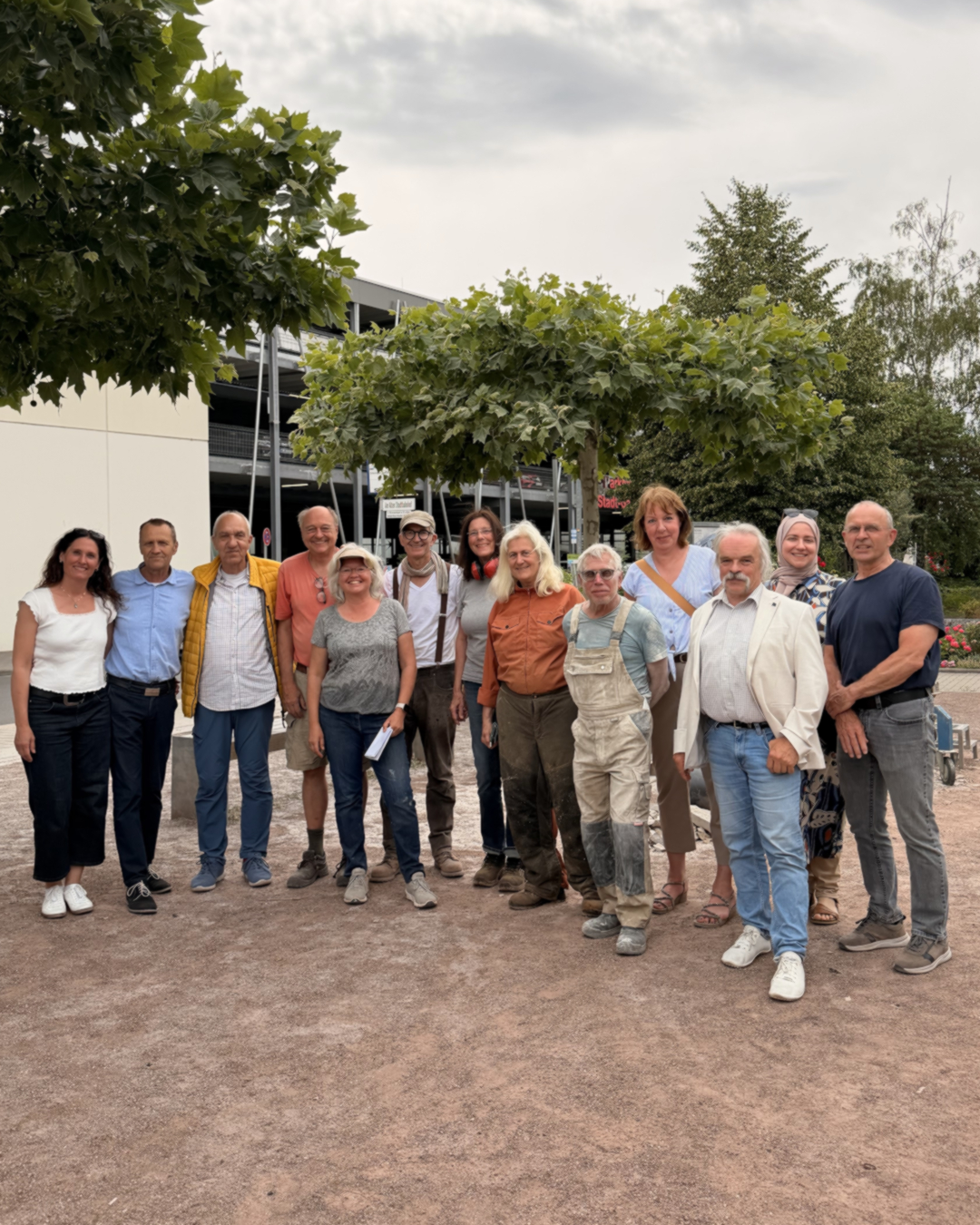 Gruppe von zwölf Personen unterschiedlichen Alters steht im Freien vor Bäumen und einem Parkhaus, lächelt in die Kamera. Eine Person trägt eine Kopfbedeckung, eine andere Arbeitskleidung. (KI-Alternativtext)