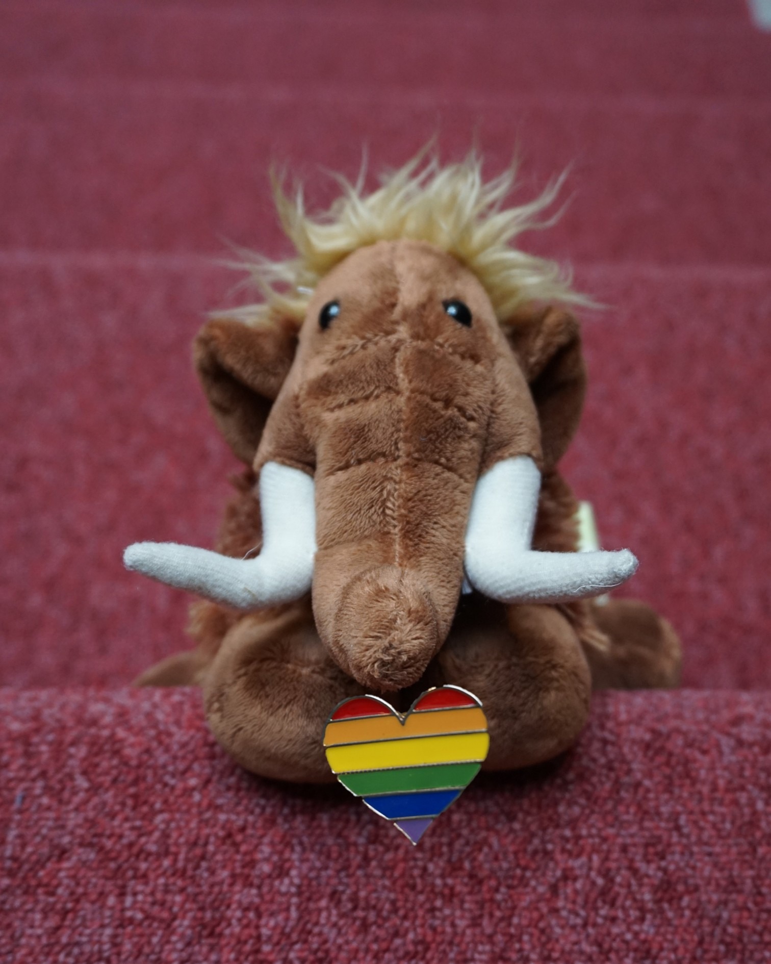 Braunes Plüschelefant mit blondem Haarschopf und weißen Stoßzähnen sitzt auf rotem Teppich, vor ihm ein Herz-Anstecker in Regenbogenfarben. (KI-Alternativtext)