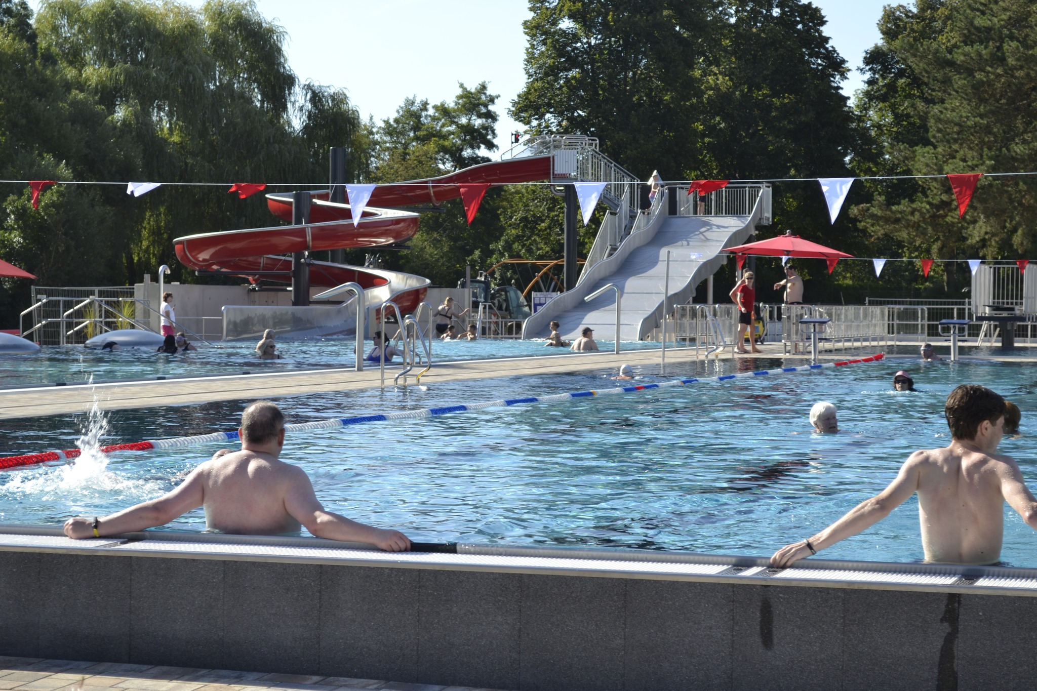 Dieser Alternativtext wurde KI-generiert: Ein Freibad mit Rutschen und verschiedenen Becken. Menschen planschen und schwimmen im Wasser, während andere am Beckenrand sitzen.