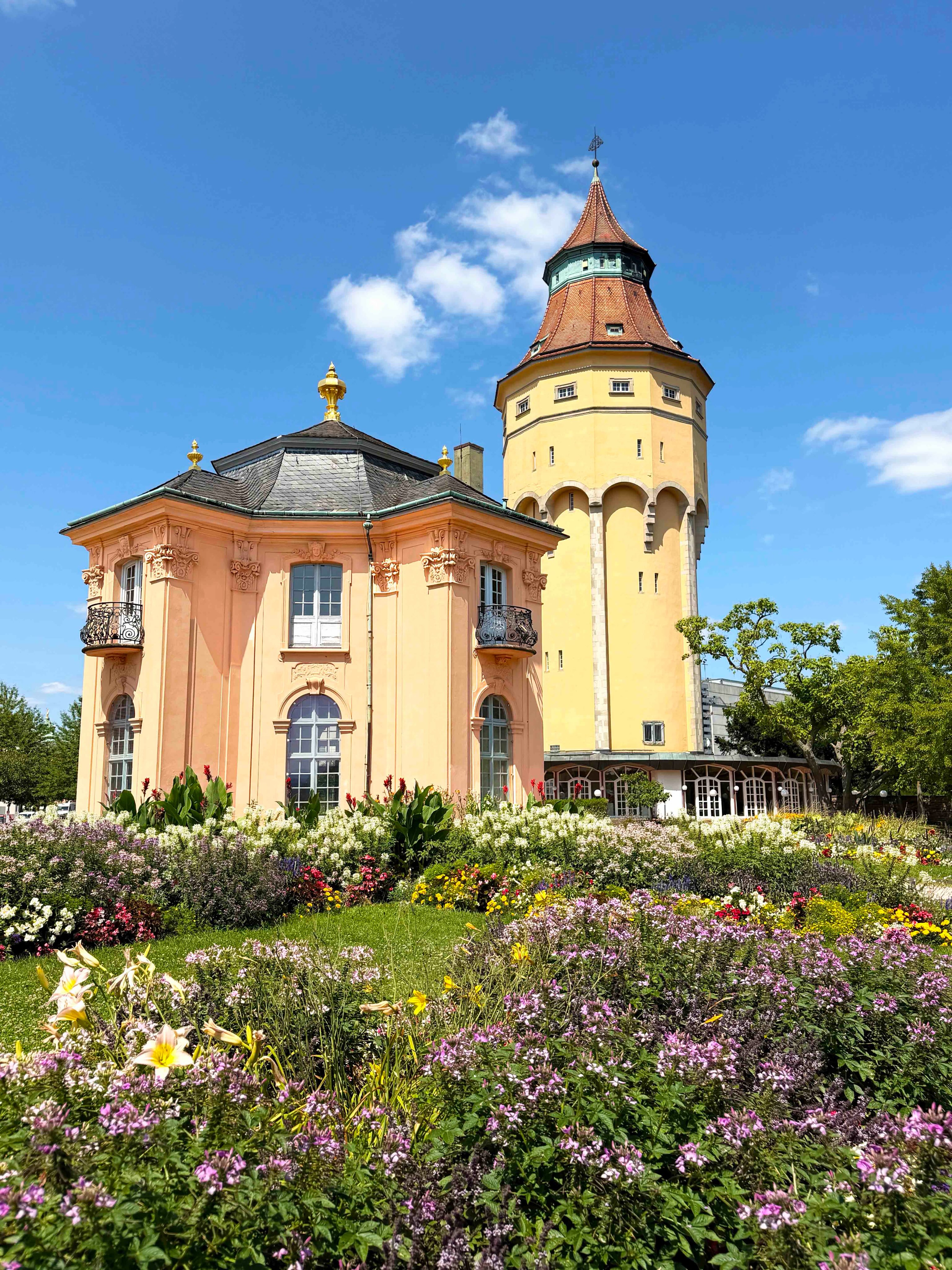 Im Vordergrund erstreckt sich ein üppiges Blumenbeet mit bunten Blüten. Dahinter stehen zwei Gebäude: links ein pfirsichfarbener Pavillon mit dunklem Dach und goldenen Verzierungen sowie rechts ein hoher, gelber Turm mit spitzem, rotbraunem Dach. Der Himmel ist blau mit vereinzelten weißen Wolken. (KI-Alternativtext)
