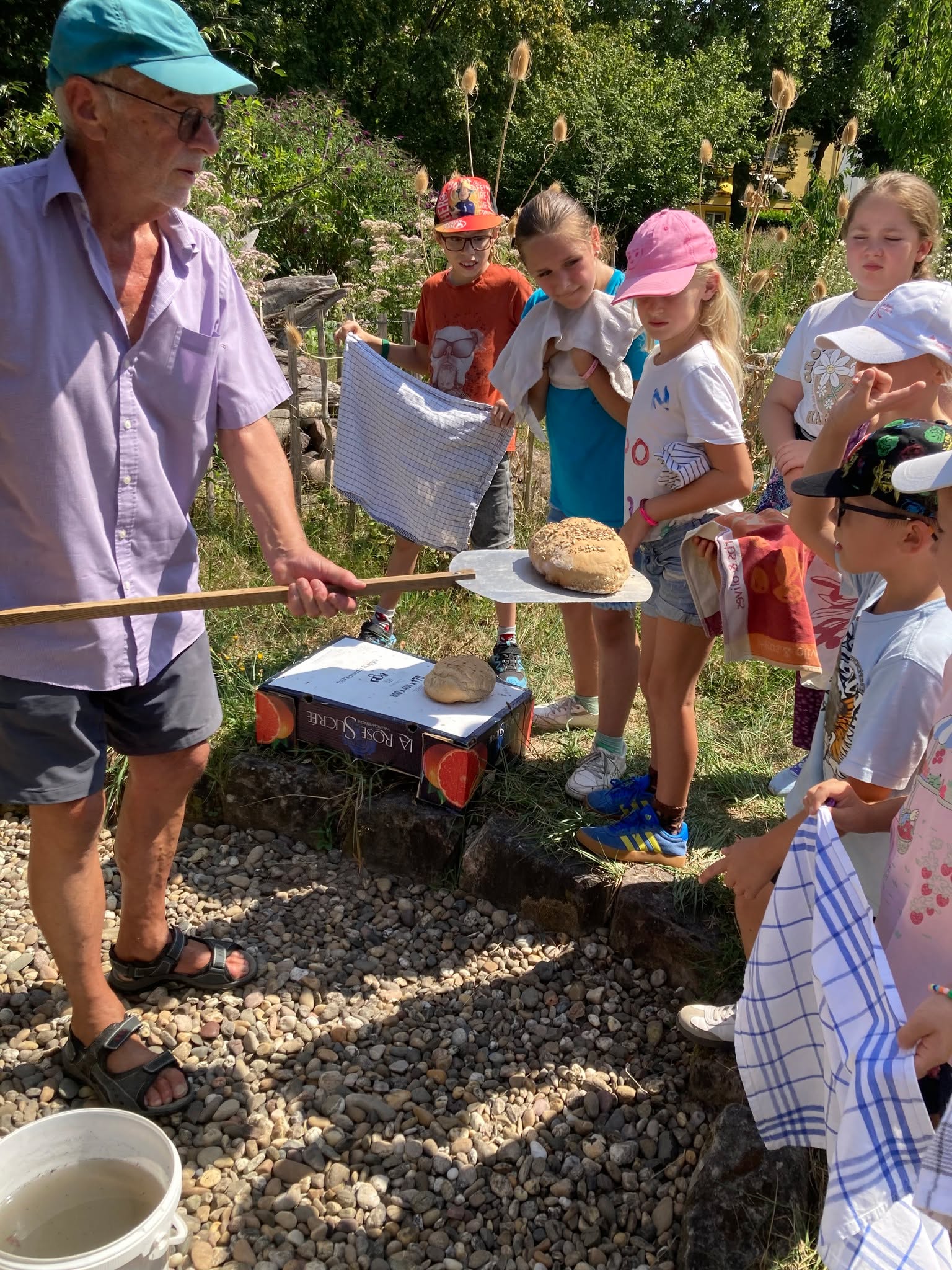Eine Person mit Kopfbedeckung und lila Hemd hält einen langen Holzlöffel mit einem frischen Brotlaib in die Höhe, während eine Gruppe von Kindern zuschaut. Auf einer Kiste mit der Aufschrift „LA ROSE SUCREE“ liegt ein weiteres Brot. Im Vordergrund steht ein weißer Eimer auf dem Kiesboden. (KI-Alternativtext)