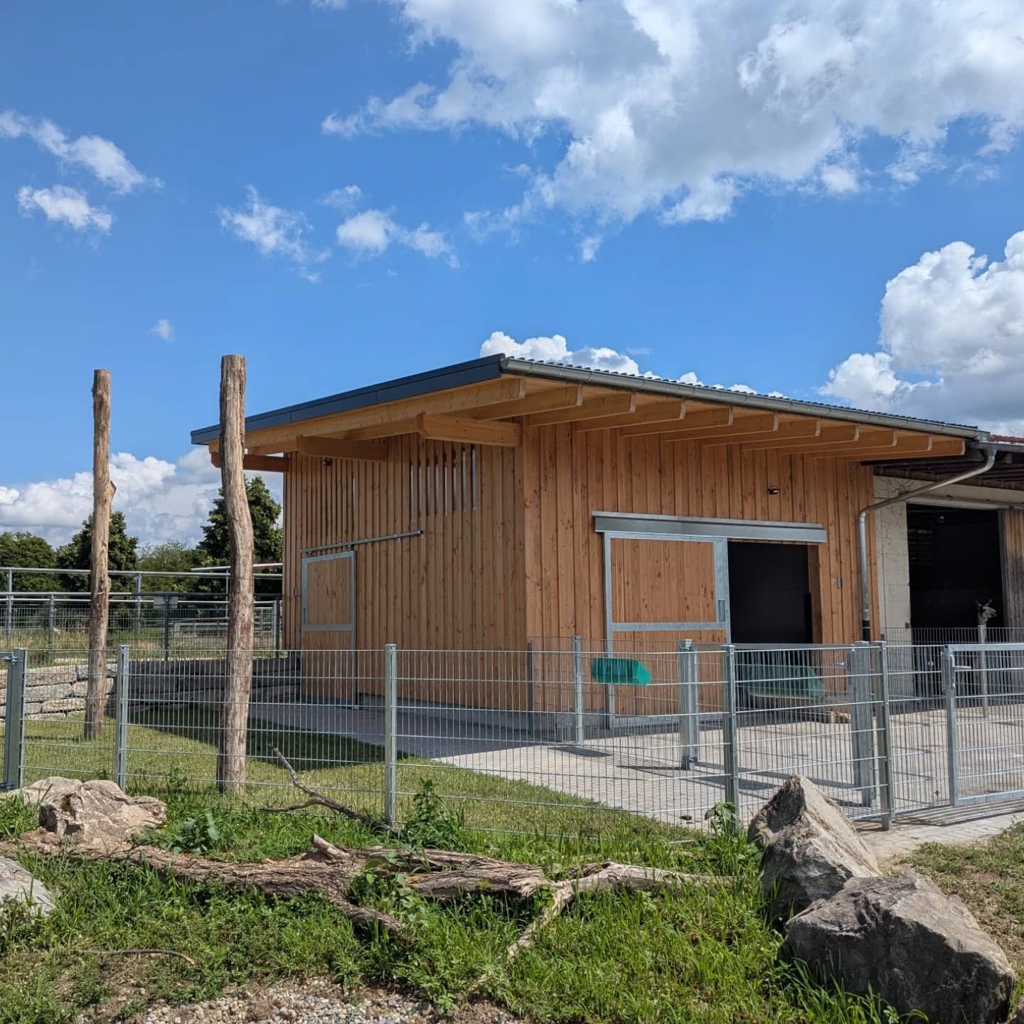 Ein gemauertes Holzgebäude mit Zaun ist im Bild zu sehen. Der Himmel ist blau mit ein paar Wolken.