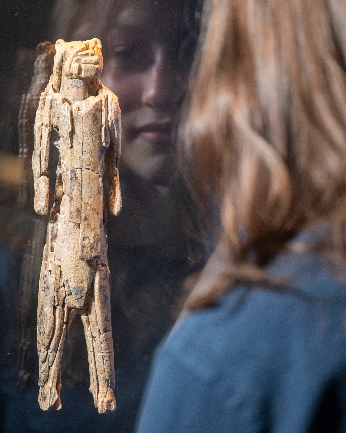 Dieser Alternativtext wurde KI-generiert: Eine Figur ist hinter Glas ausgestellt. Im Hintergrund ist eine verschwommene Person mit langen Haaren zu sehen.