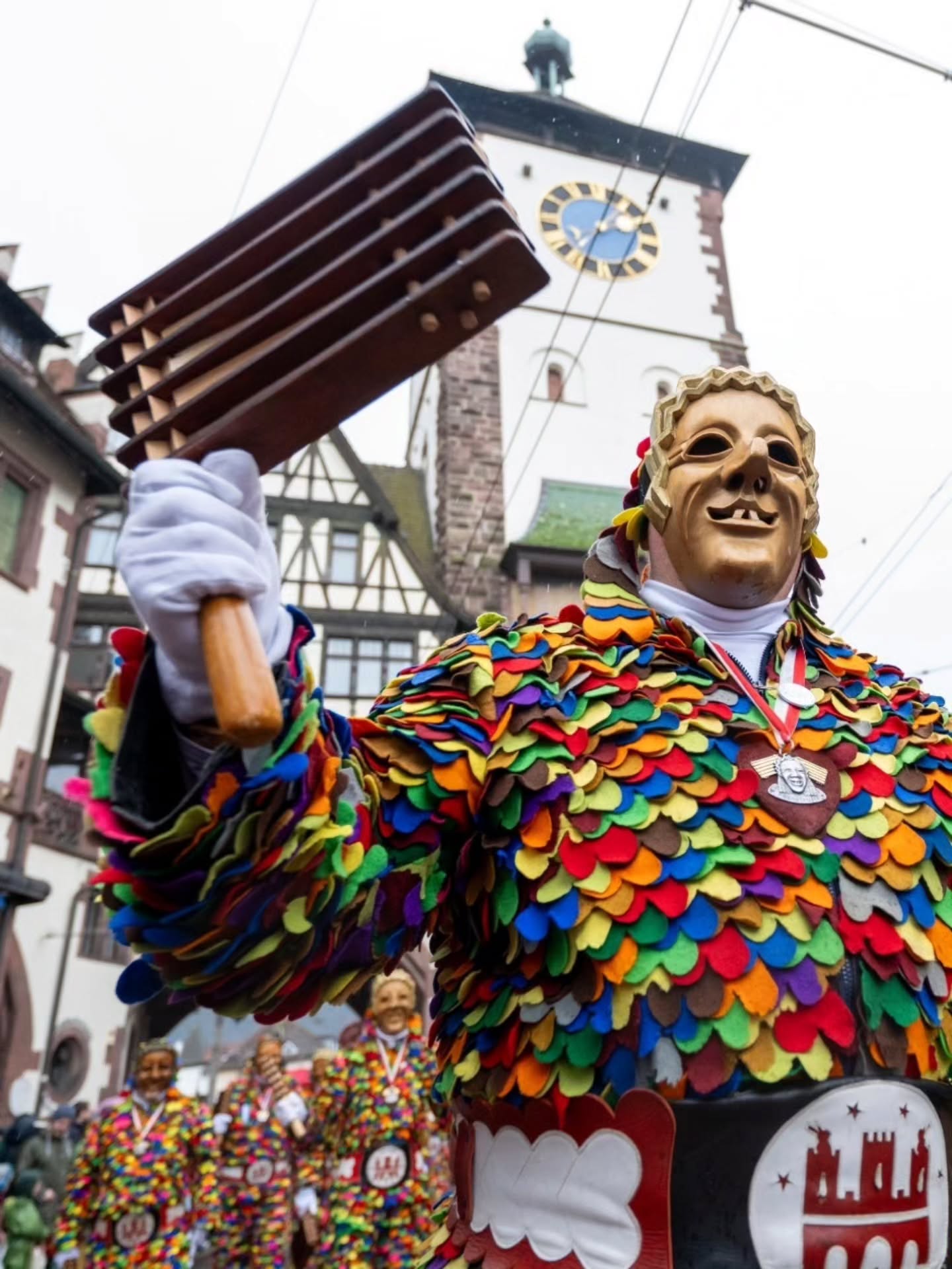Dieser Alternativtext wurde KI-generiert: Ein Mensch in einem farbenfrohen Kostüm und einer Maske hält ein Musikinstrument hoch. Im Hintergrund ist eine Stadtansicht mit einem Turm zu sehen.
