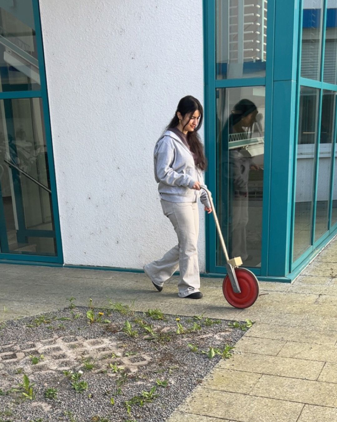 Dieser Alternativtext wurde KI-generiert: Eine Person fährt auf einem Tretroller neben einem Gebäude mit großen Fenstern. Das Bild ist farbig und zeigt einen hellen Innenhof.