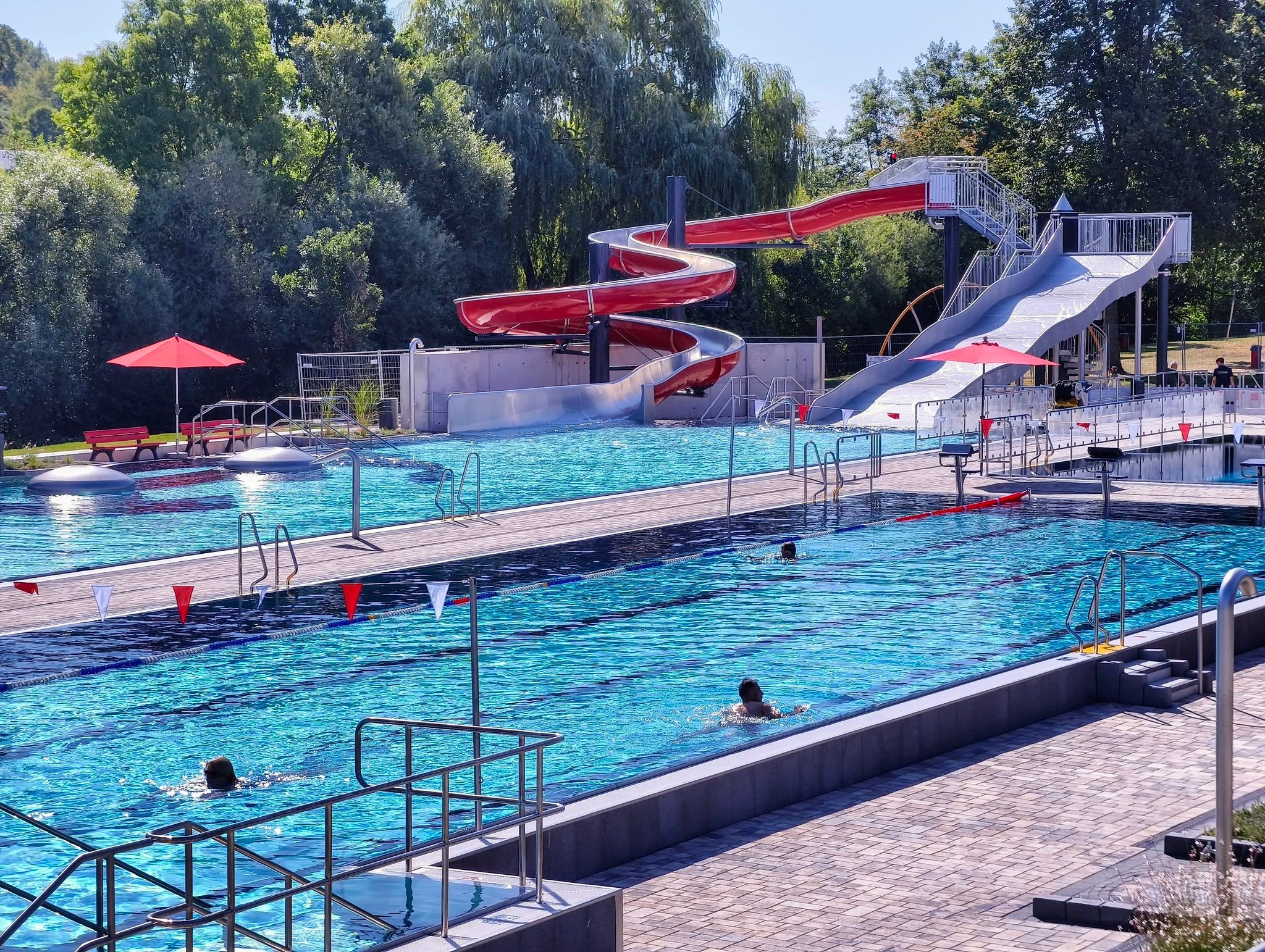 Ein sonniges Freibad mit einem großen Schwimmbecken im Vordergrund, in dem mehrere Personen schwimmen. Im Hintergrund stehen zwei große Wasserrutschen, eine rote Spirale und eine weiße, vor einer dichten Baumreihe. (KI-Alternativtext)